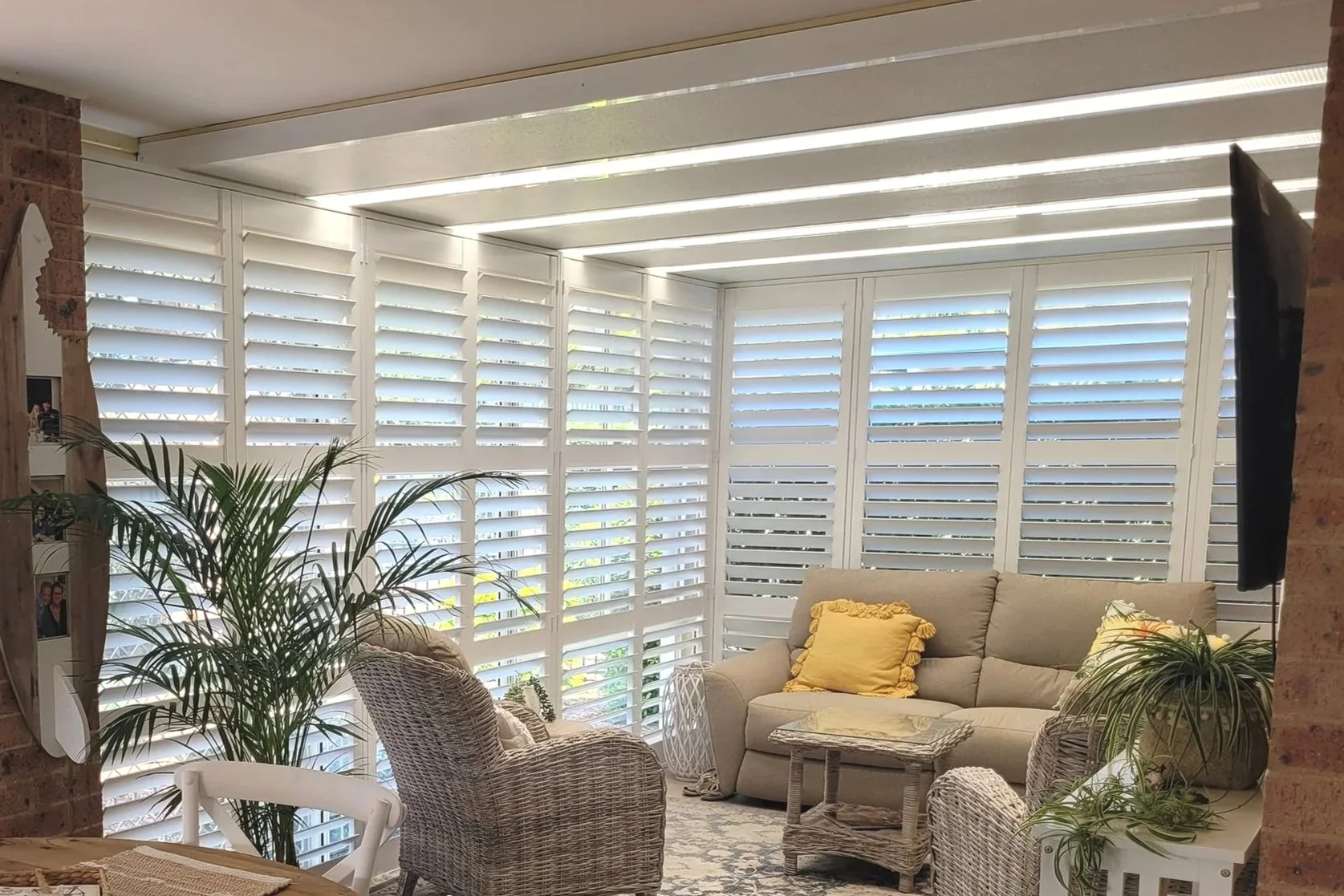 Living room with large white window shutters, beige sofa with yellow pillows, wicker chairs, indoor plants, and a wall-mounted TV.