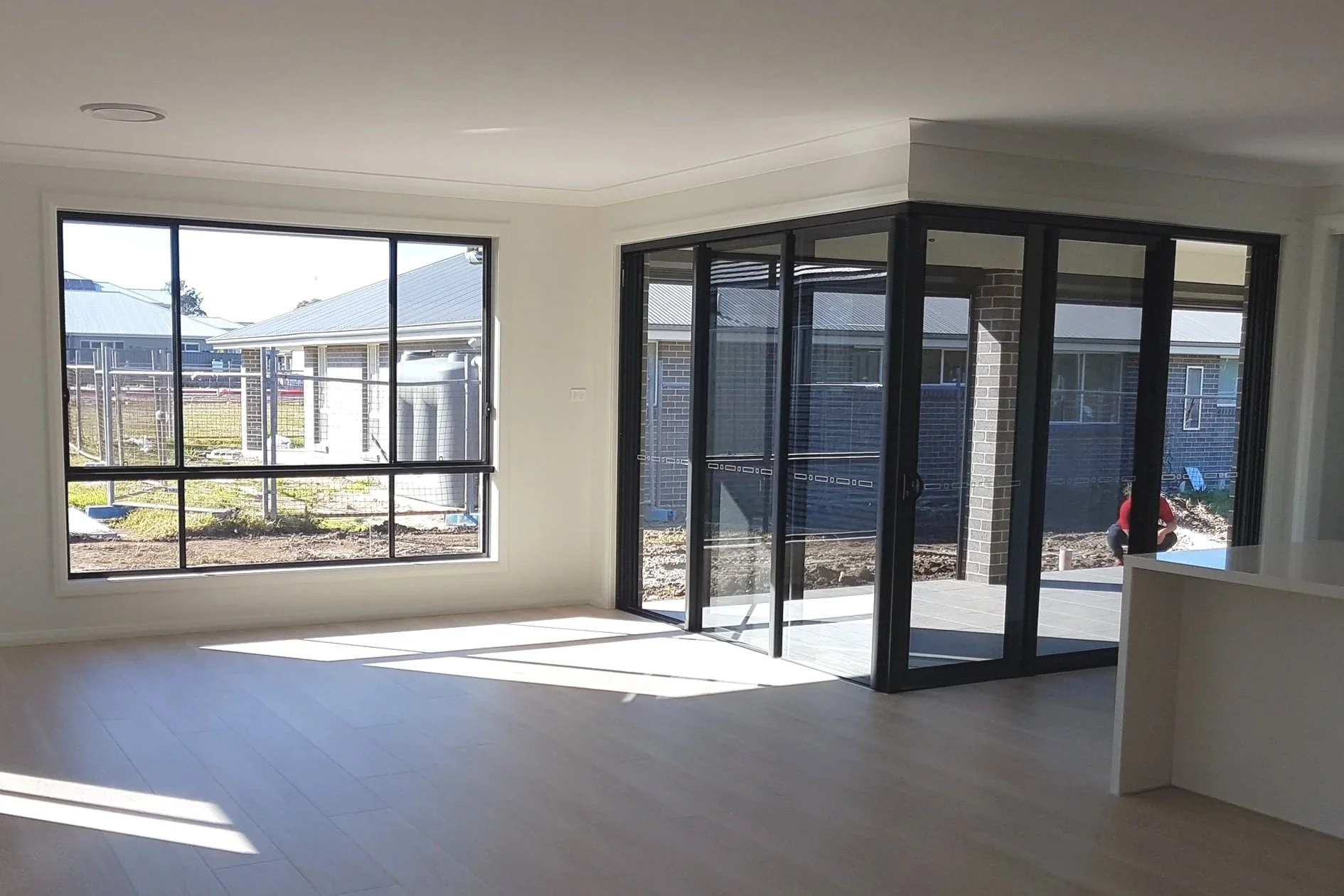 Interior of a new house during construction, featuring large windows, a sliding glass door, and an outdoor patio area with a person working in the background.