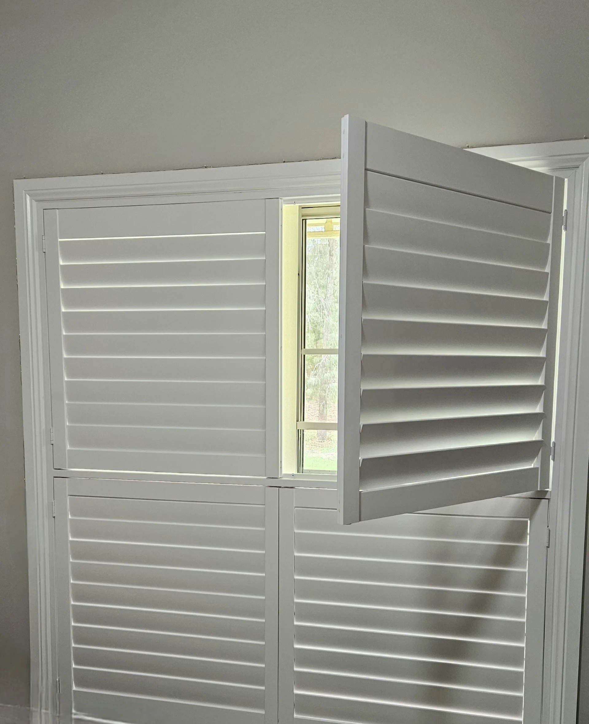 White plantation shutters partially open in front of a window, with a view of trees outside.