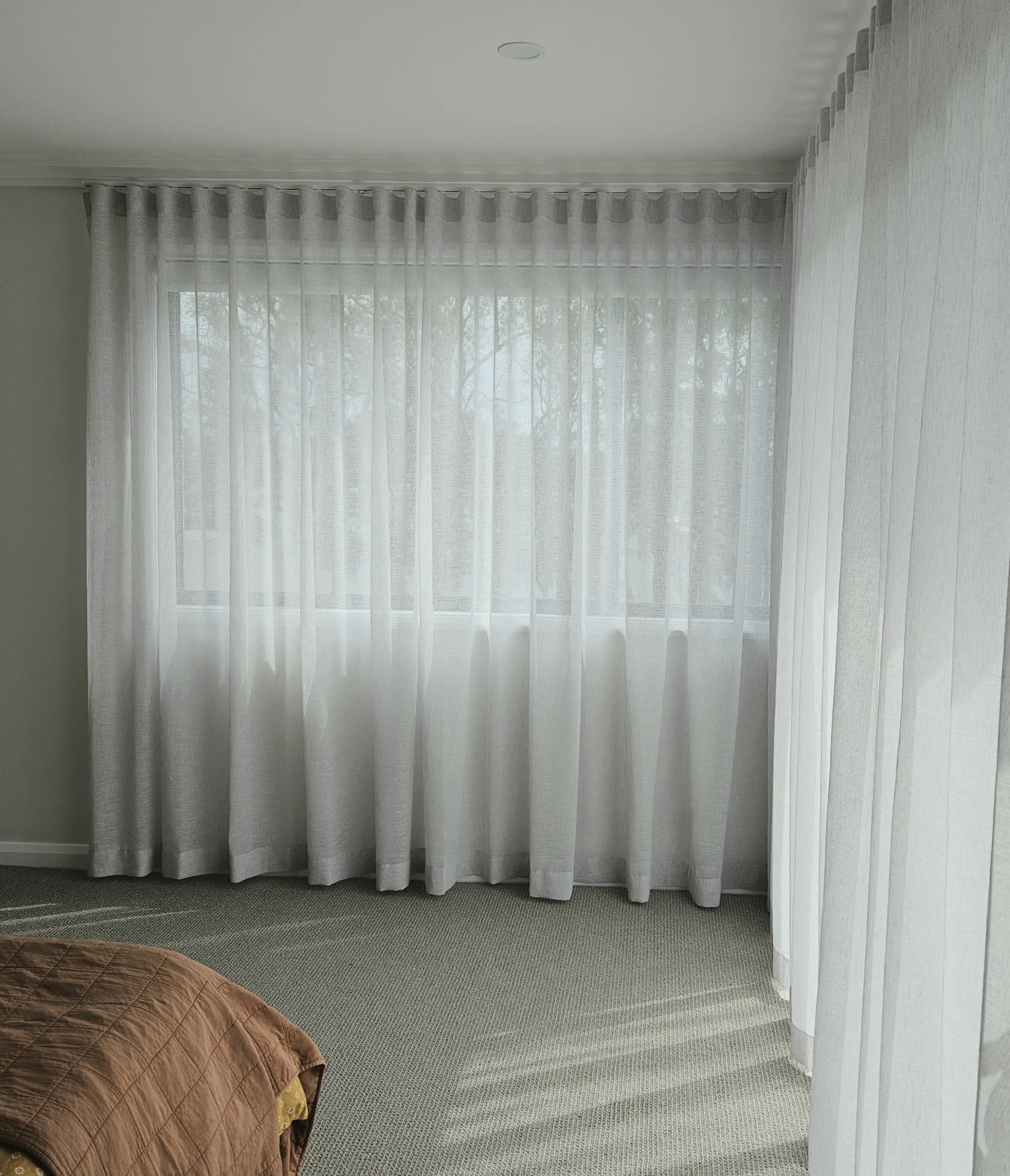 Living room with beige carpet, light green walls, sheer white curtains on a window, part of a bed with a brown quilted cover in the foreground.