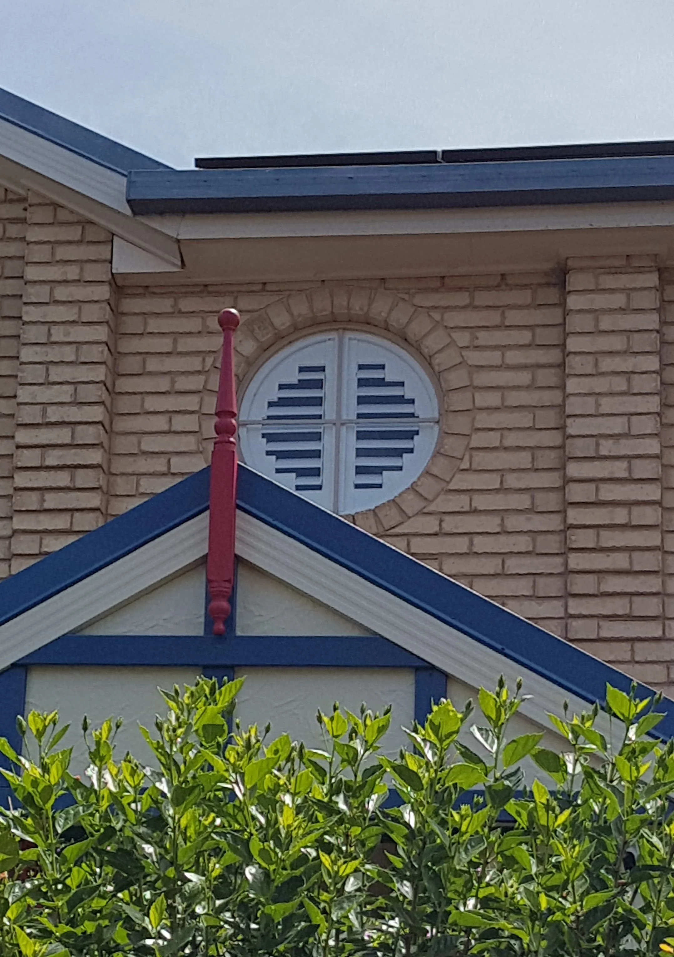 Close-up of a house with a yellow brick exterior, a blue roof with decorative red trim, and a round window with shutters above a hedge of green bushes.