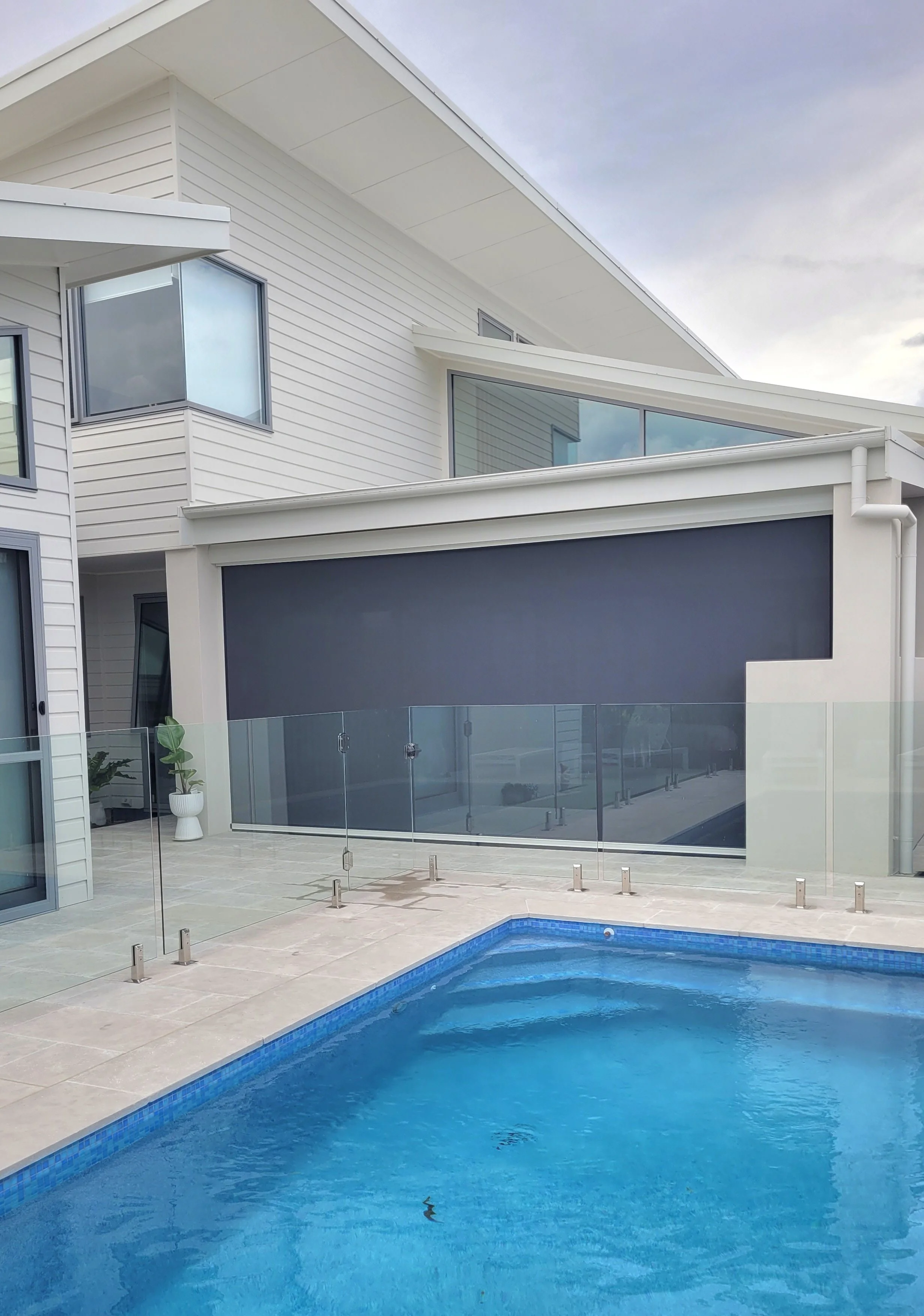 Modern house with white siding and large windows, overlooking a swimming pool with a glass fence around it, on a cloudy day.