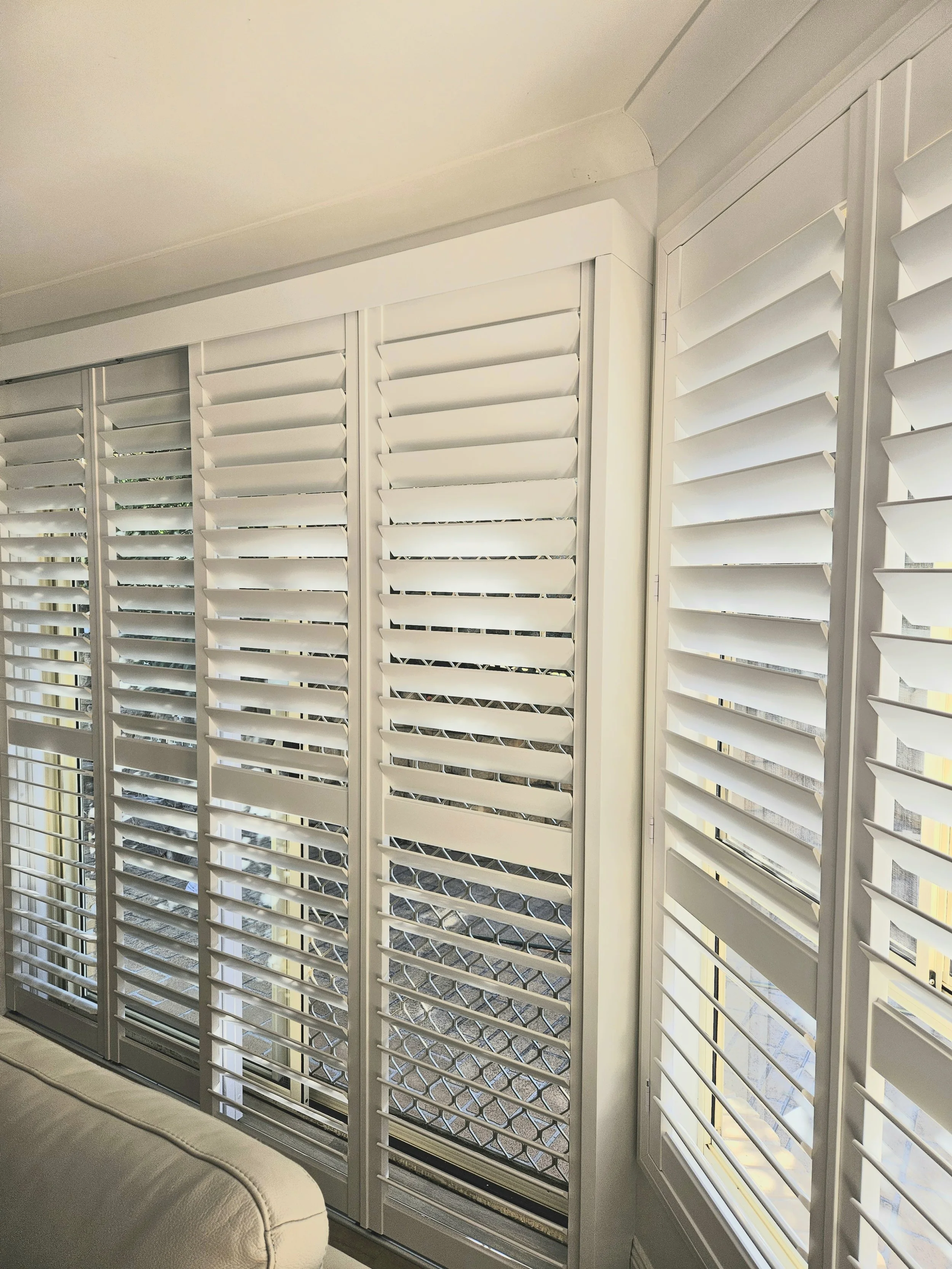White plantation shutters on large windows in a bright room, with a view of a backyard.