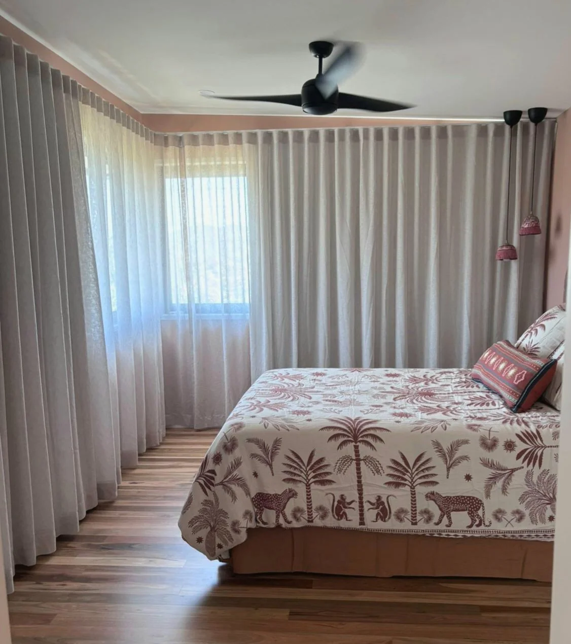 Bedroom with a bed covered in a tropical tree and animal patterned bedspread, sheer white curtains, a ceiling fan, and two hanging pink lamps.