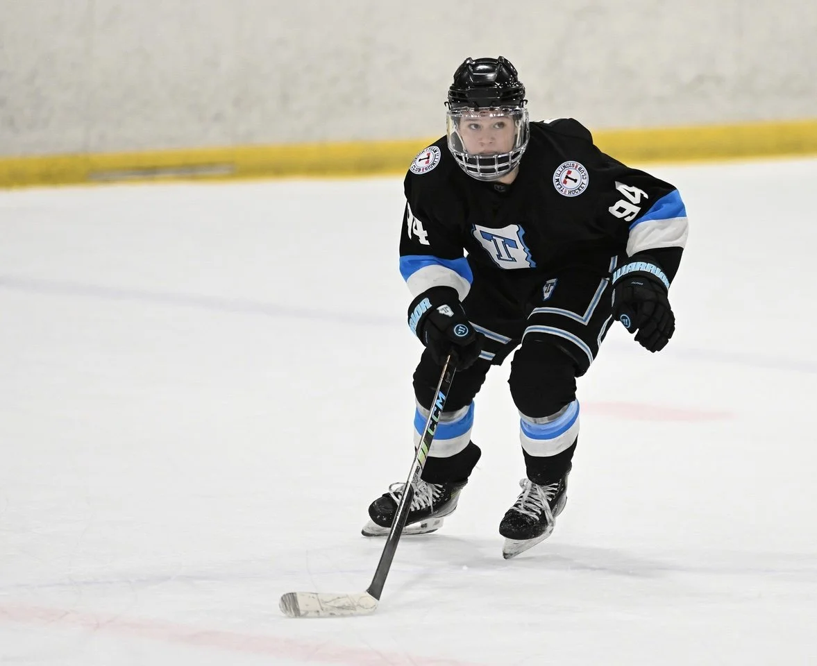 A young ice hockey player in black and blue uniform skating on ice with a hockey stick.