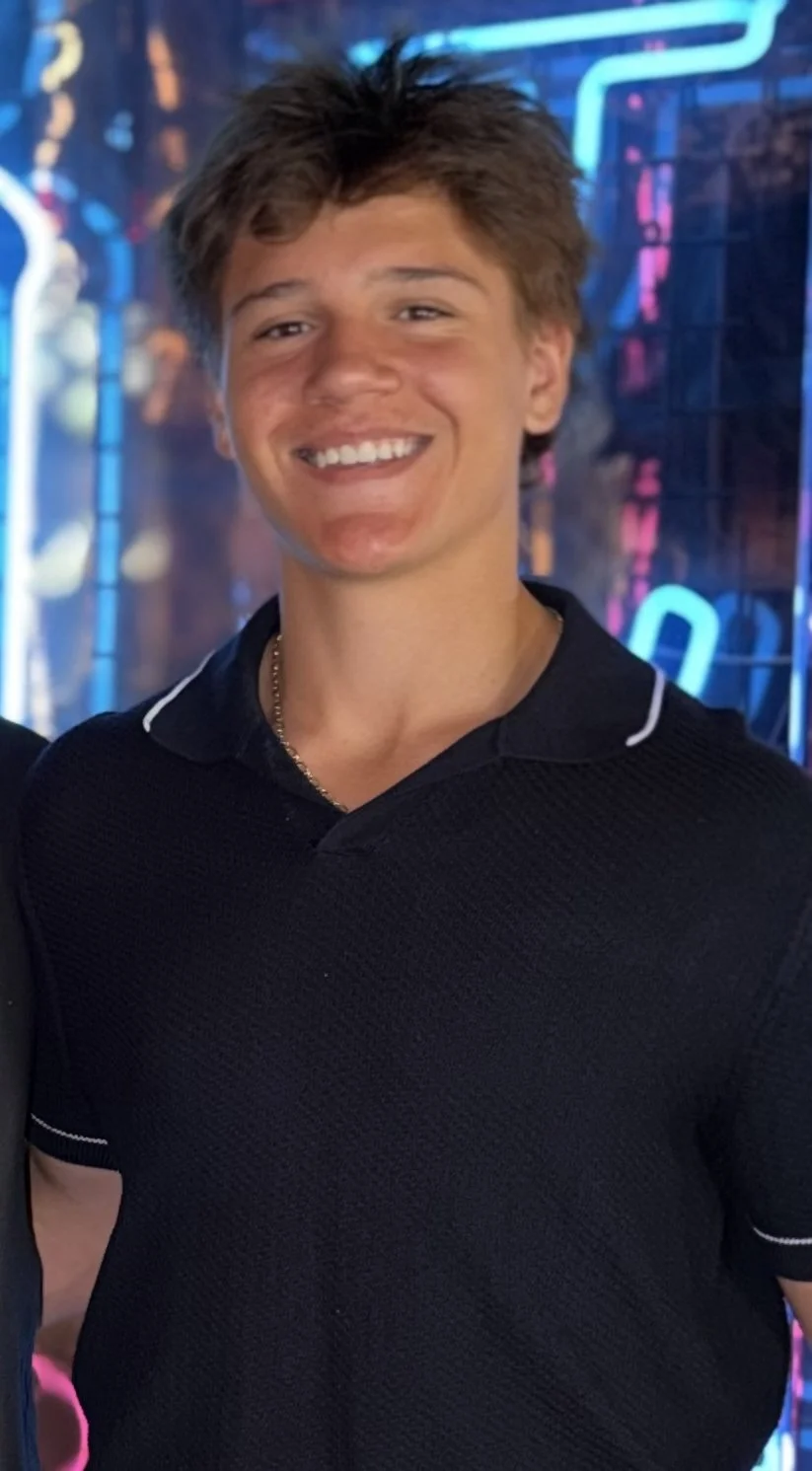 A young man with brown hair, smiling, wearing a black polo shirt with white piping, standing in front of colorful neon lights.