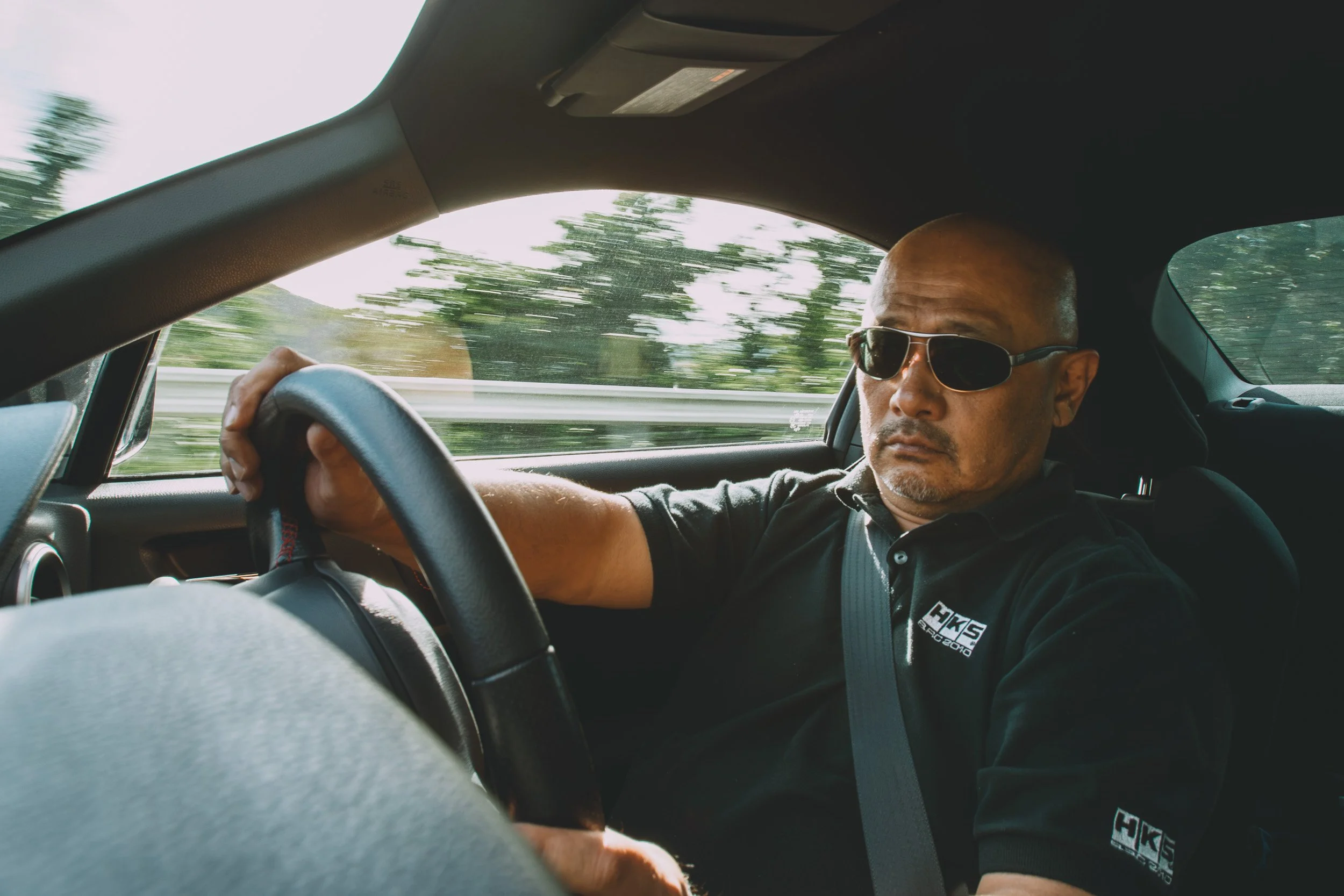 Masanori Kuwabara wearing an HKS-branded polo shirt gripping the steering wheel while driving, motion blur outside the window.