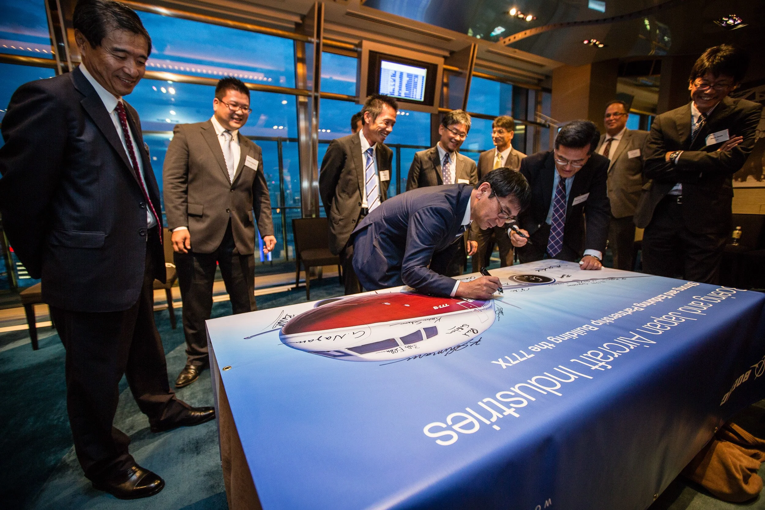 Executive signing a Boeing 777X partnership banner as colleagues look on, Tokyo skyline in the background