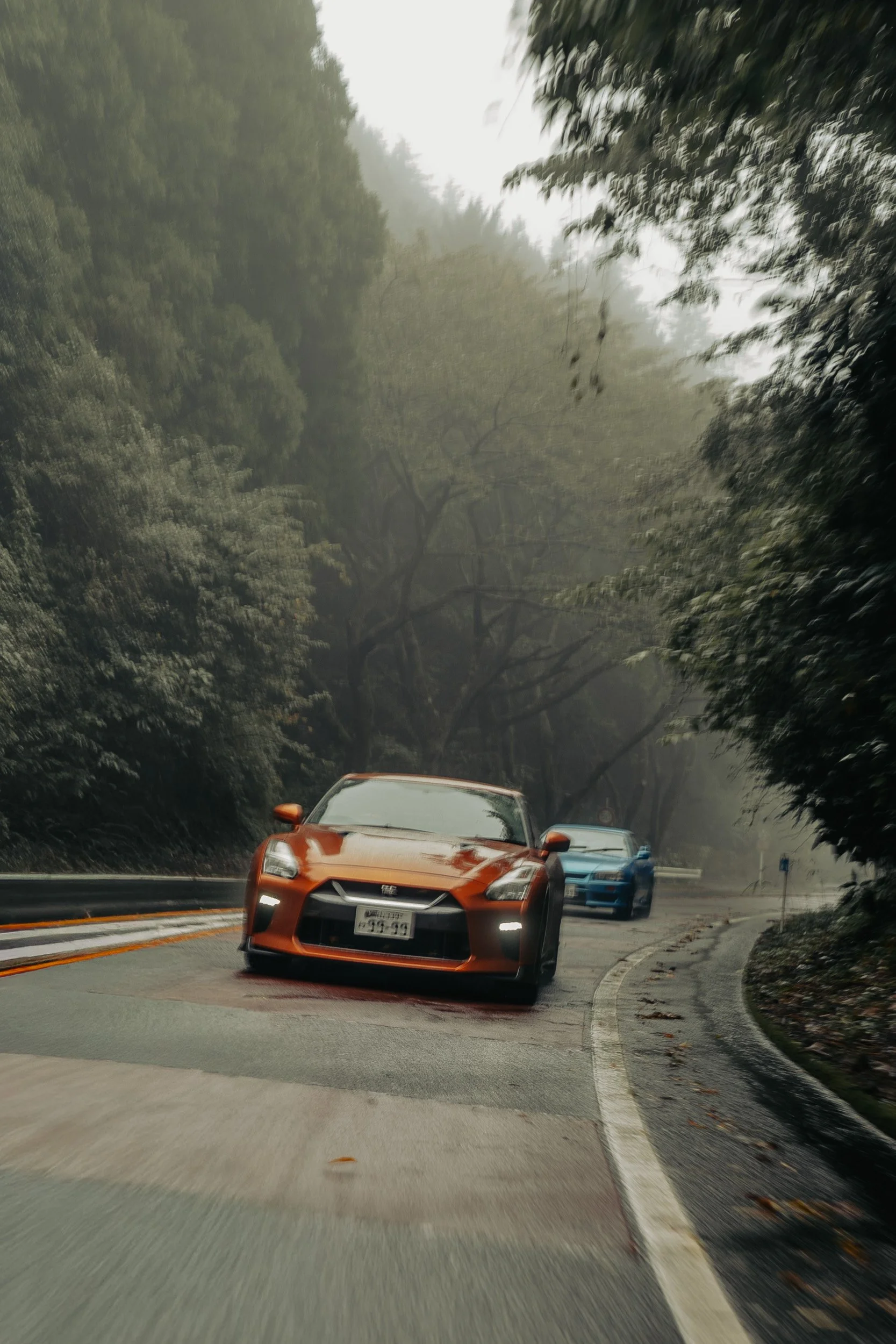 Orange Nissan R35 GT-R leading a blue R34 Skyline GT-R through the mist on the Hakone Turnpike, Japan