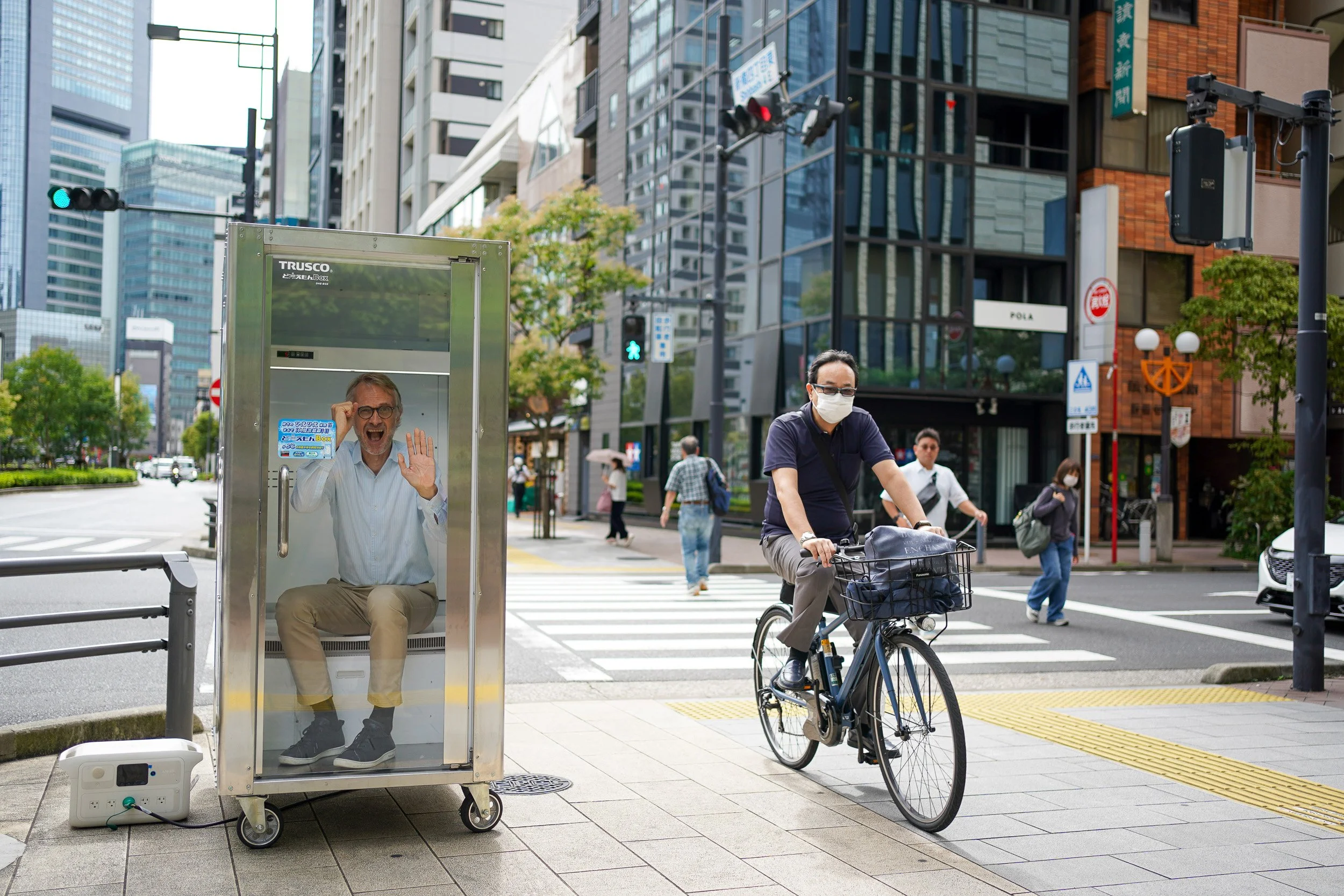 Asia editor of The Times and The Sunday Times, Richard Lloyd Parry, reacts from inside a portable cooling booth on a Tokyo street corner as a masked cyclist rides past during the summer heat.