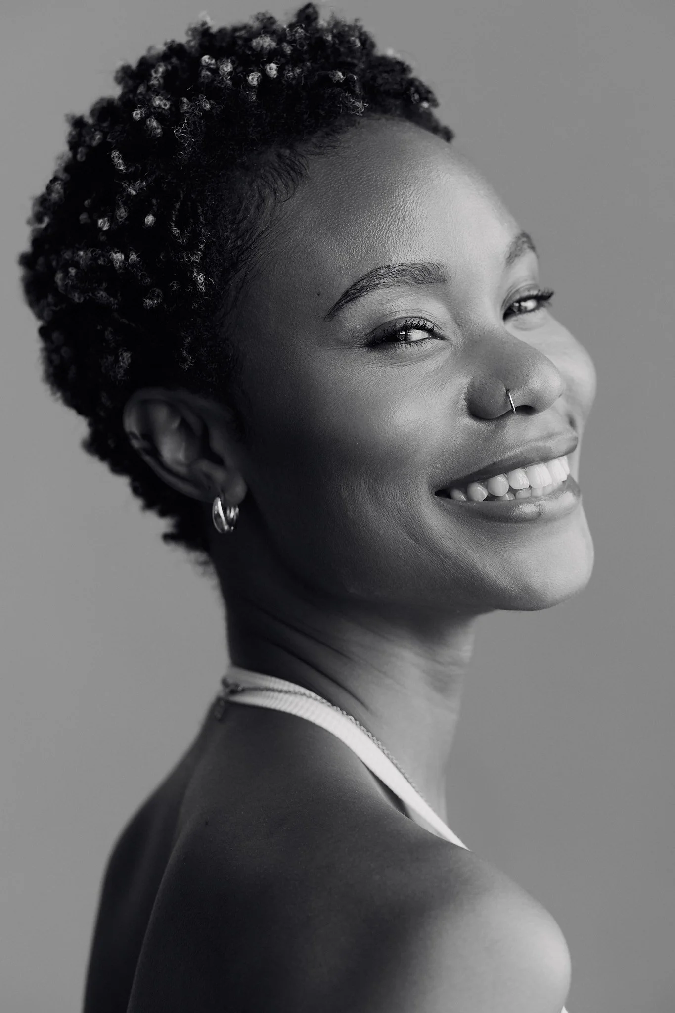 A black and white portrait of a smiling woman with short curly hair, wearing hoop earrings, a nose ring, a choker necklace, and a tank top, looking slightly to the side.