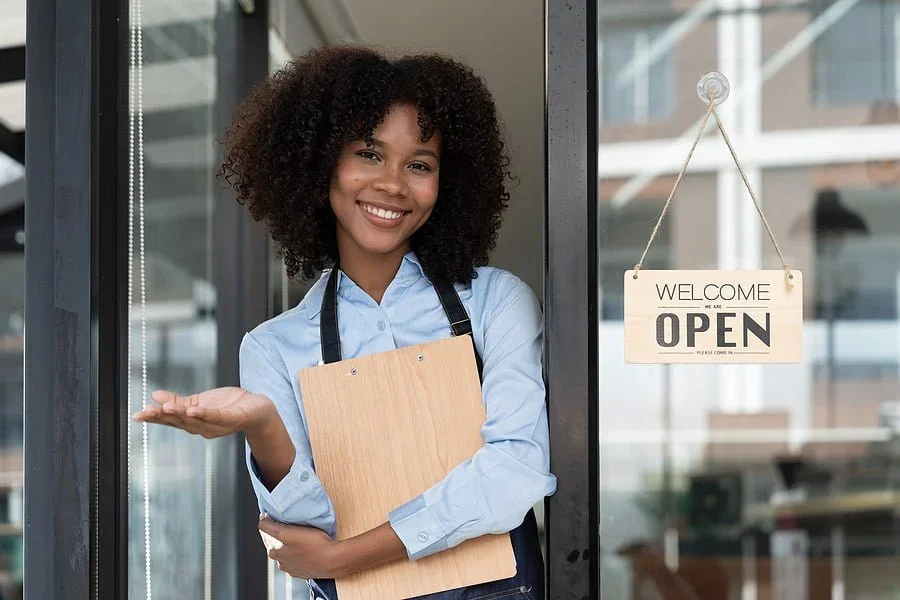 Smiling woman standing in the doorway of a cafe or shop, holding a menu or clipboard, with an 'Open' sign hanging outside.