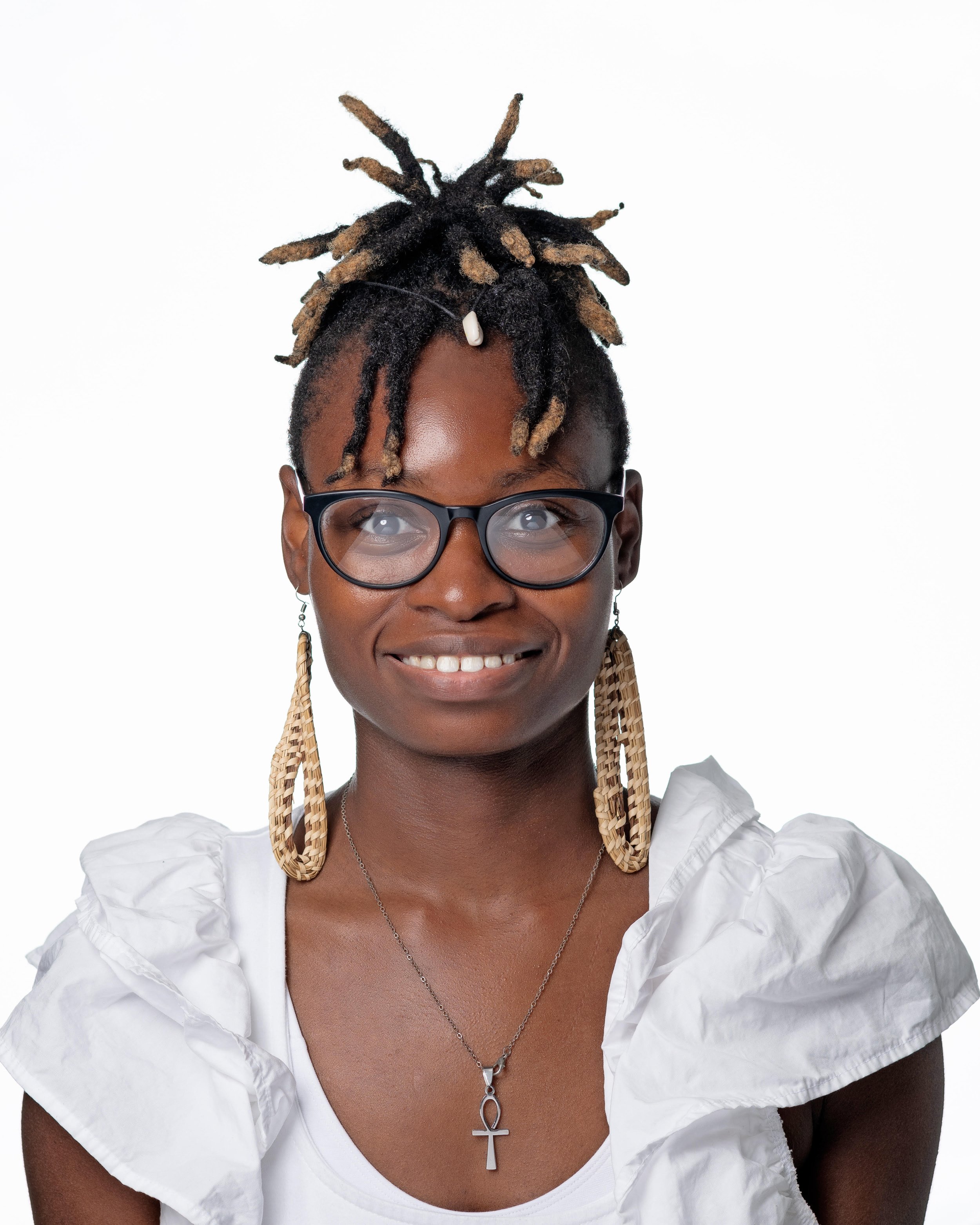 A woman with dark skin, wearing glasses with black frames, smiling slightly, and dressed in a white blouse featuring puffed sleeves and ruffles. She has a silver necklace with an ankh pendant and long, braided earrings. Her hair is styled in dreadlocks with some tied up on top of her head.