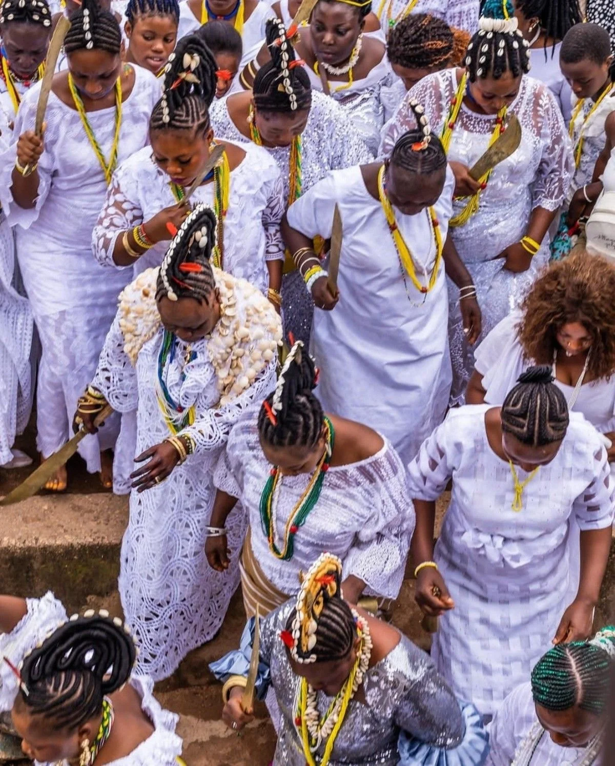 A group of women and girls dressed in traditional white garments during a cultural or ceremonial event, with intricate jewelry and hairstyles, holding sticks or staffs.