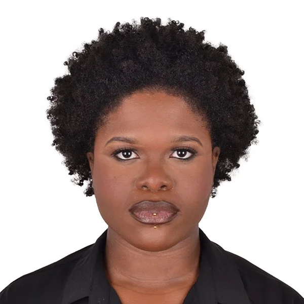 A close-up portrait of a woman with dark skin and short curly black hair, wearing a black top, against a plain white background.