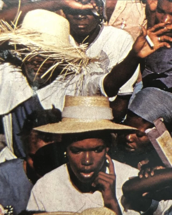 Young woman wearing a wide-brimmed straw hat with a ribbon, in a crowded outdoor setting.
