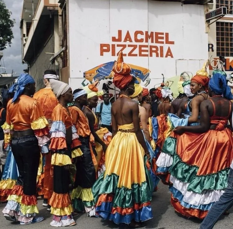 Group of people in colorful traditional dresses and headwraps, gathered on the street in front of a pizzeria sign.