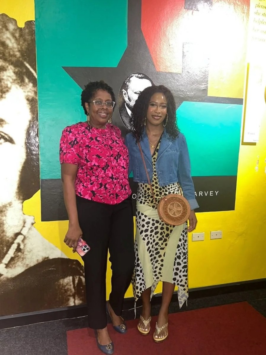 Two women standing in front of a colorful mural with a black and white photo of a man, smiling at the camera. One woman is wearing a pink floral blouse and black pants, the other wearing a denim jacket over a polka dot skirt, holding a round woven bag.