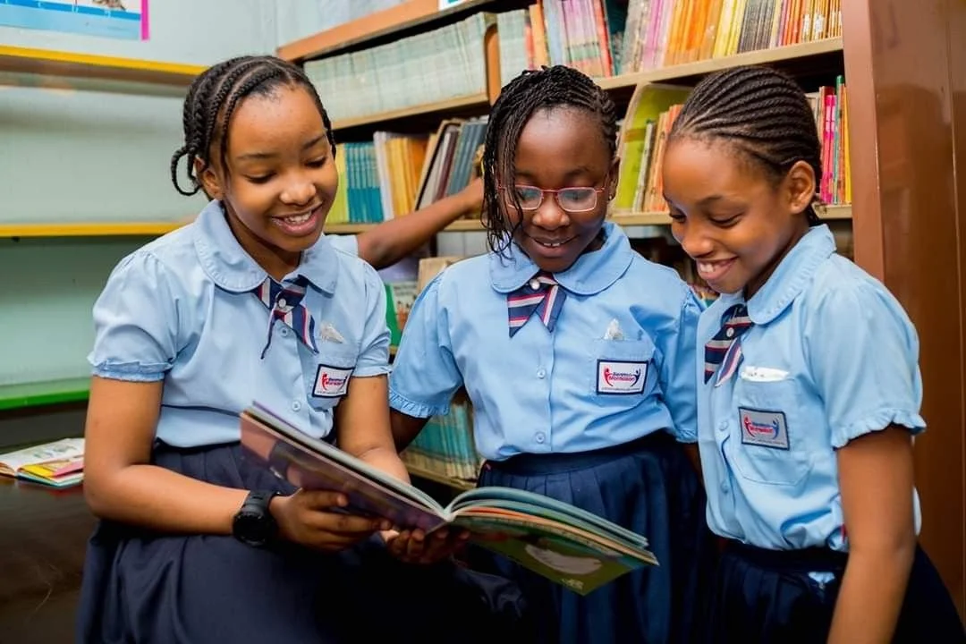 Three young girls in school uniforms smiling and looking at a book in a school library.