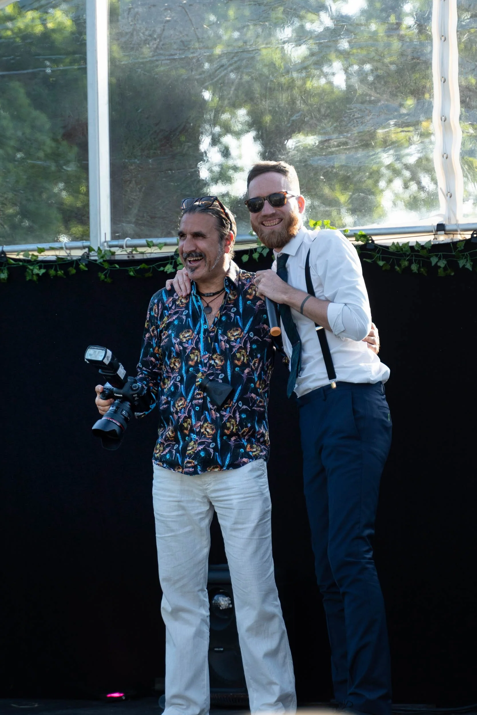 Two men smiling and embracing each other outdoors, one holding a camera, in front of a black backdrop with greenery and a transparent canopy above.