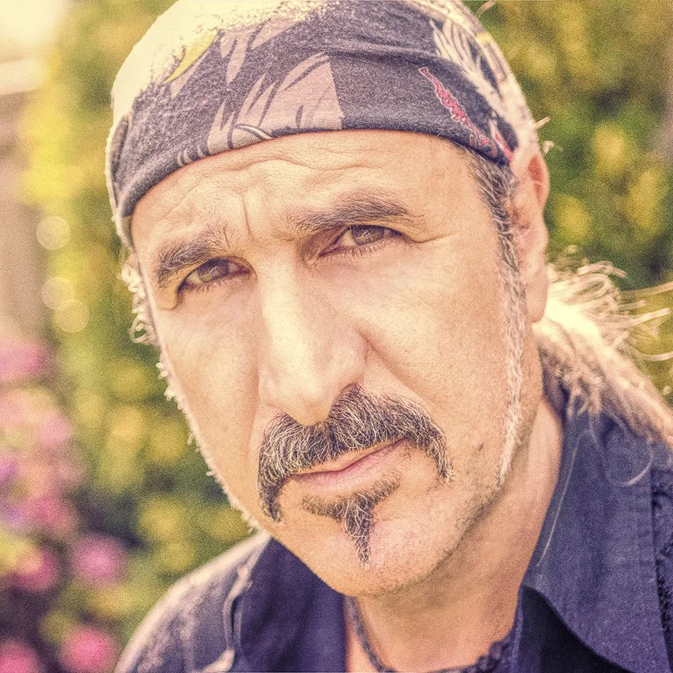 Close-up of a man with a bandana on his head, sporting a mustache and goatee, looking directly at the camera with a serious expression, with blurred greenery and pink flowers in the background.