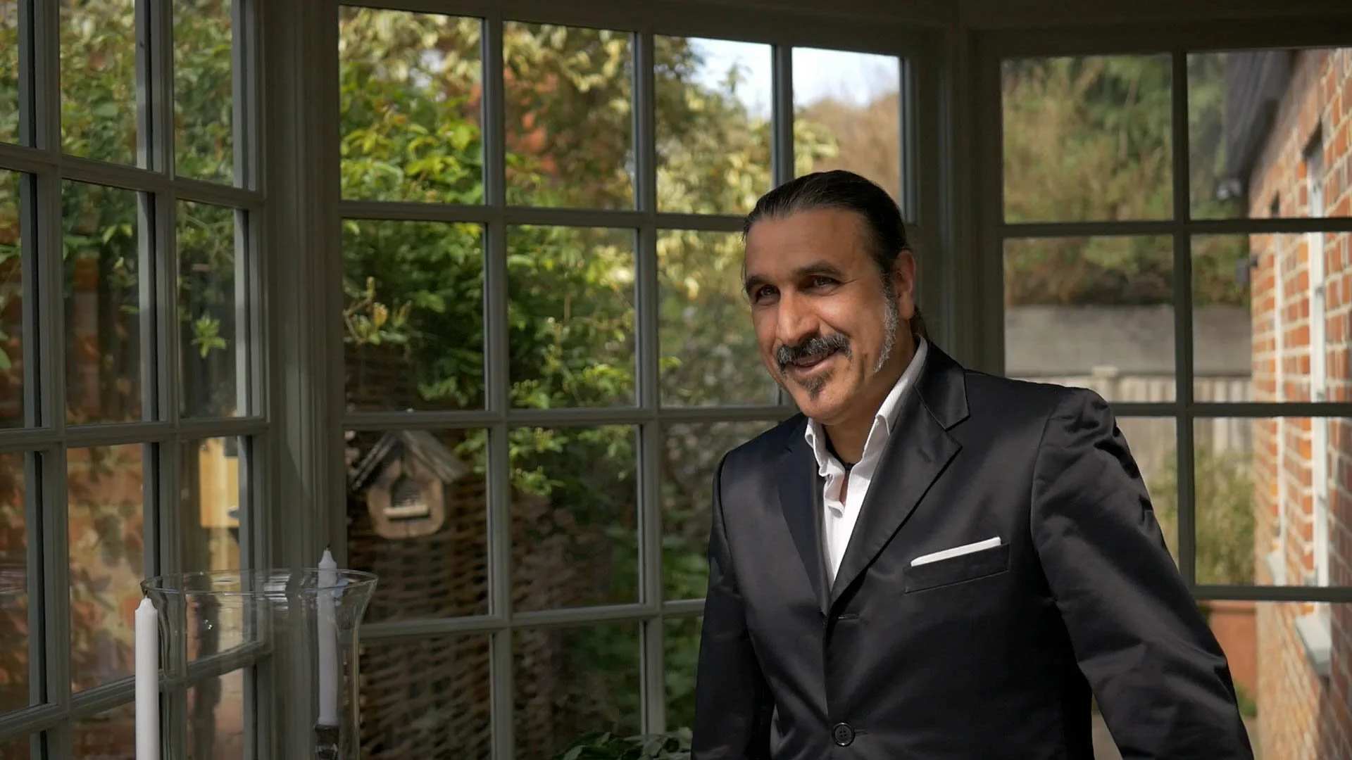 A man in a black suit smiling indoors near a window with a garden view.