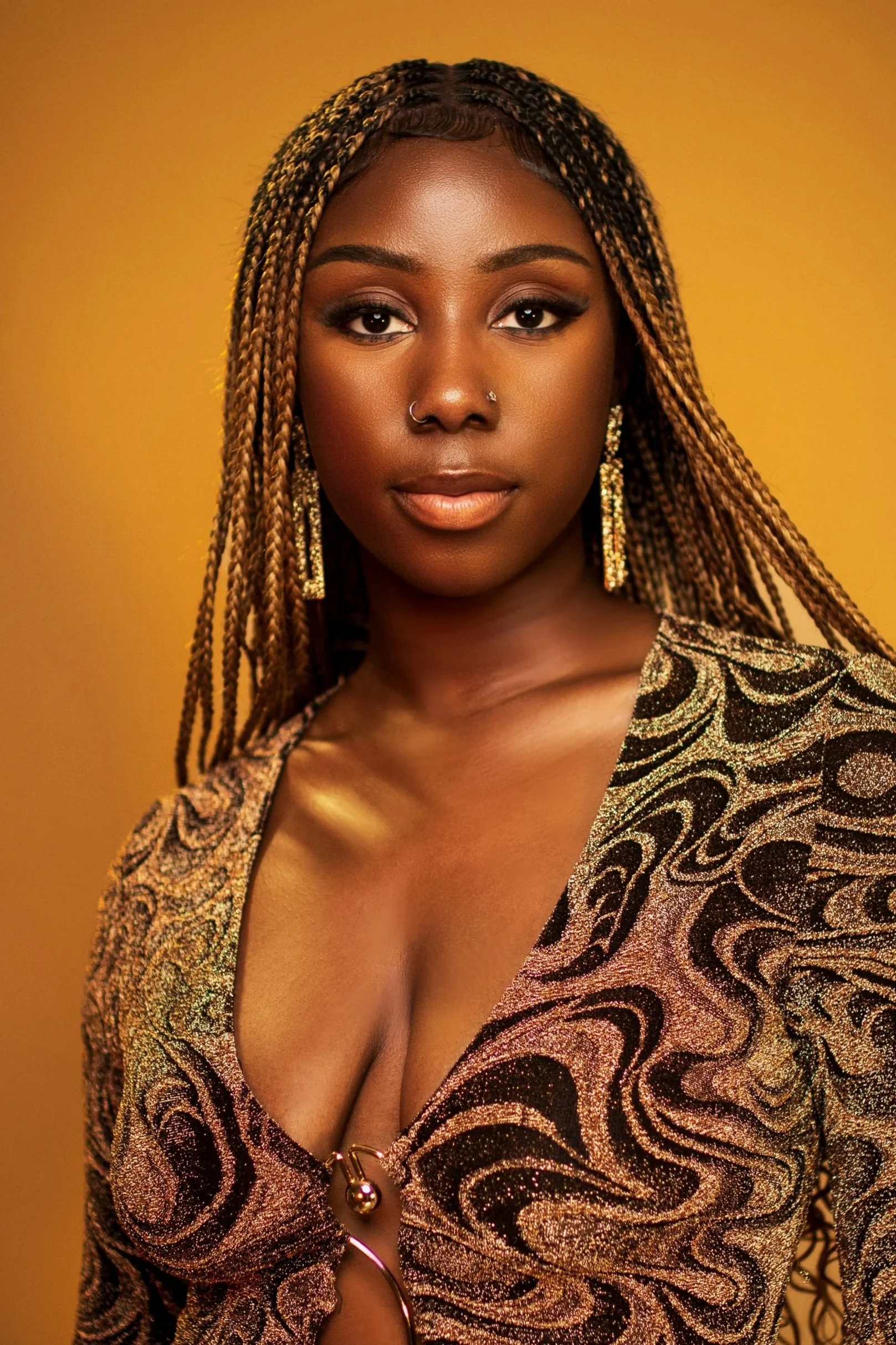 A close-up portrait of a woman with dark skin, long braided hair, and gold earrings, wearing a black and gold patterned dress with a plunging neckline, against a gold background.