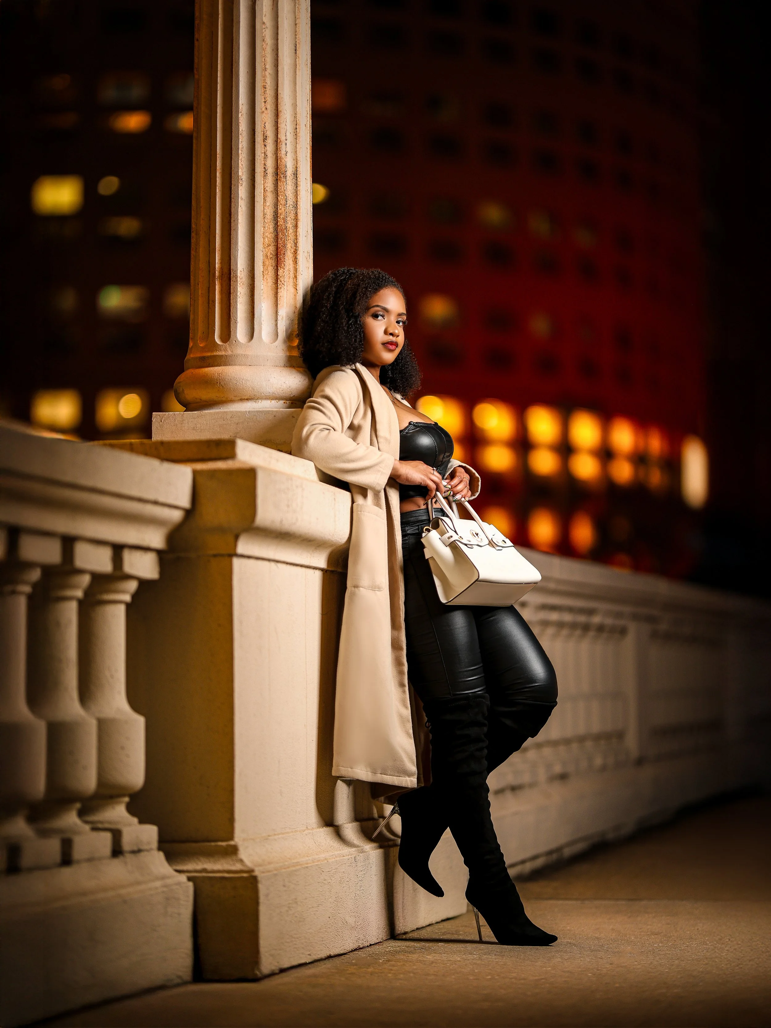 A woman with curly hair, dressed in black leather top and black pants, leaning against a stone balcony railing at night.