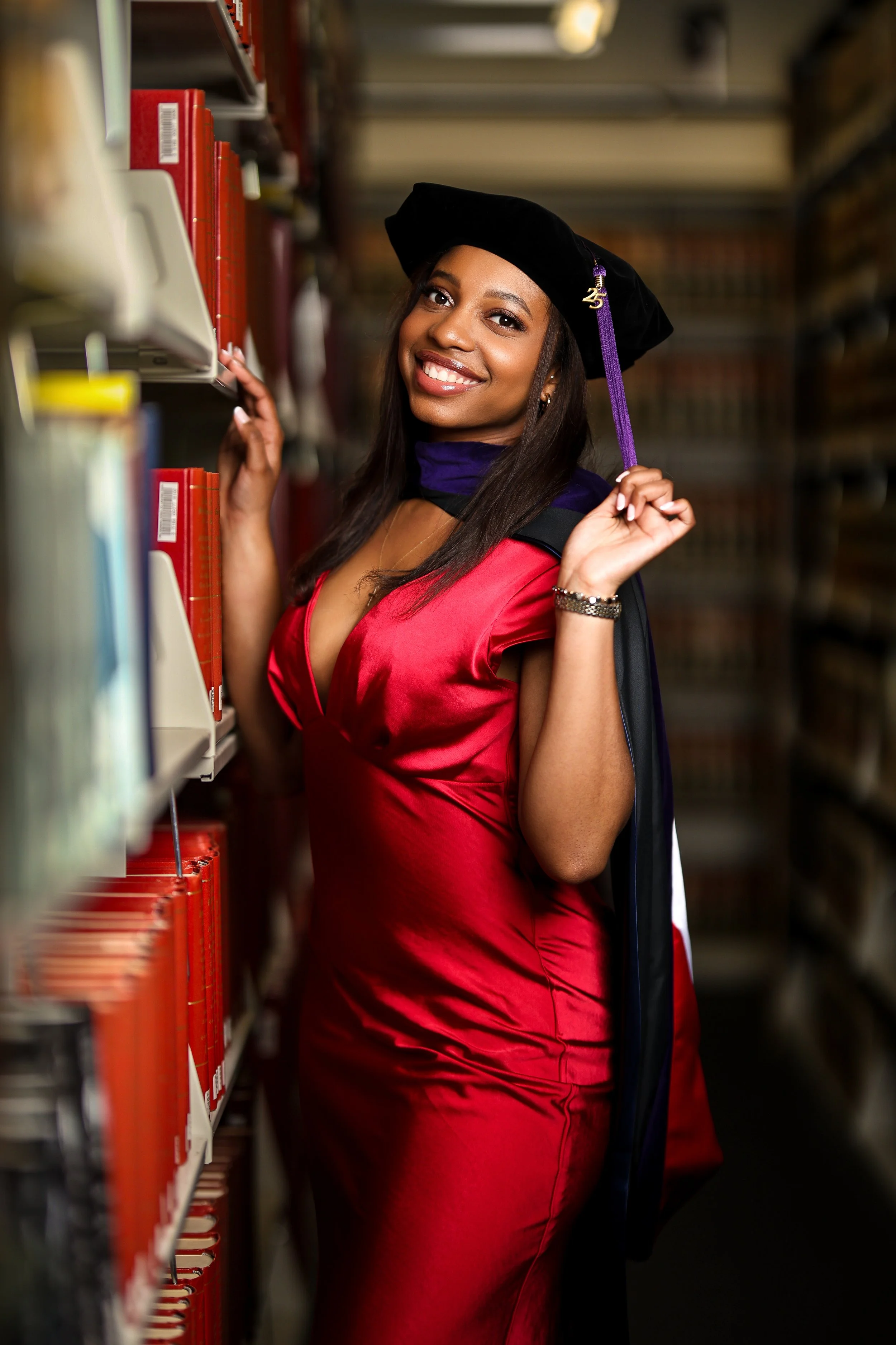 A young woman in a red graduation gown and black cap with purple tassel stands in a library aisle, smiling and looking at the camera while holding a book on a shelf.