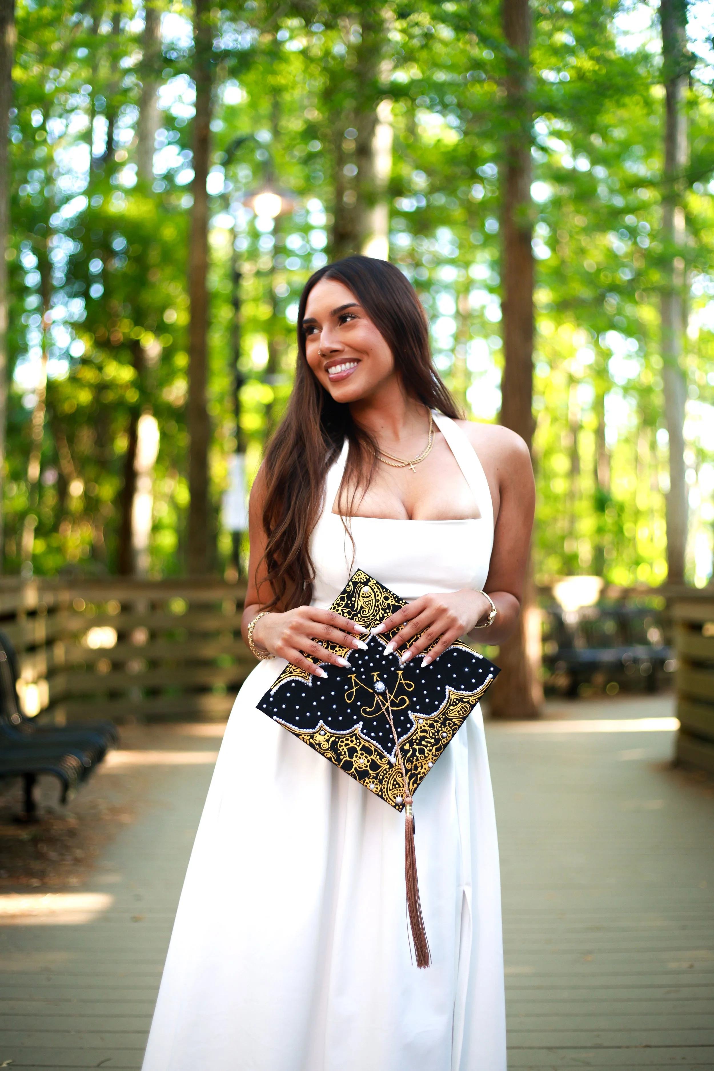 A young woman in a white dress holding a decorated graduation cap, standing outdoors in a wooded area with trees and a pathway.