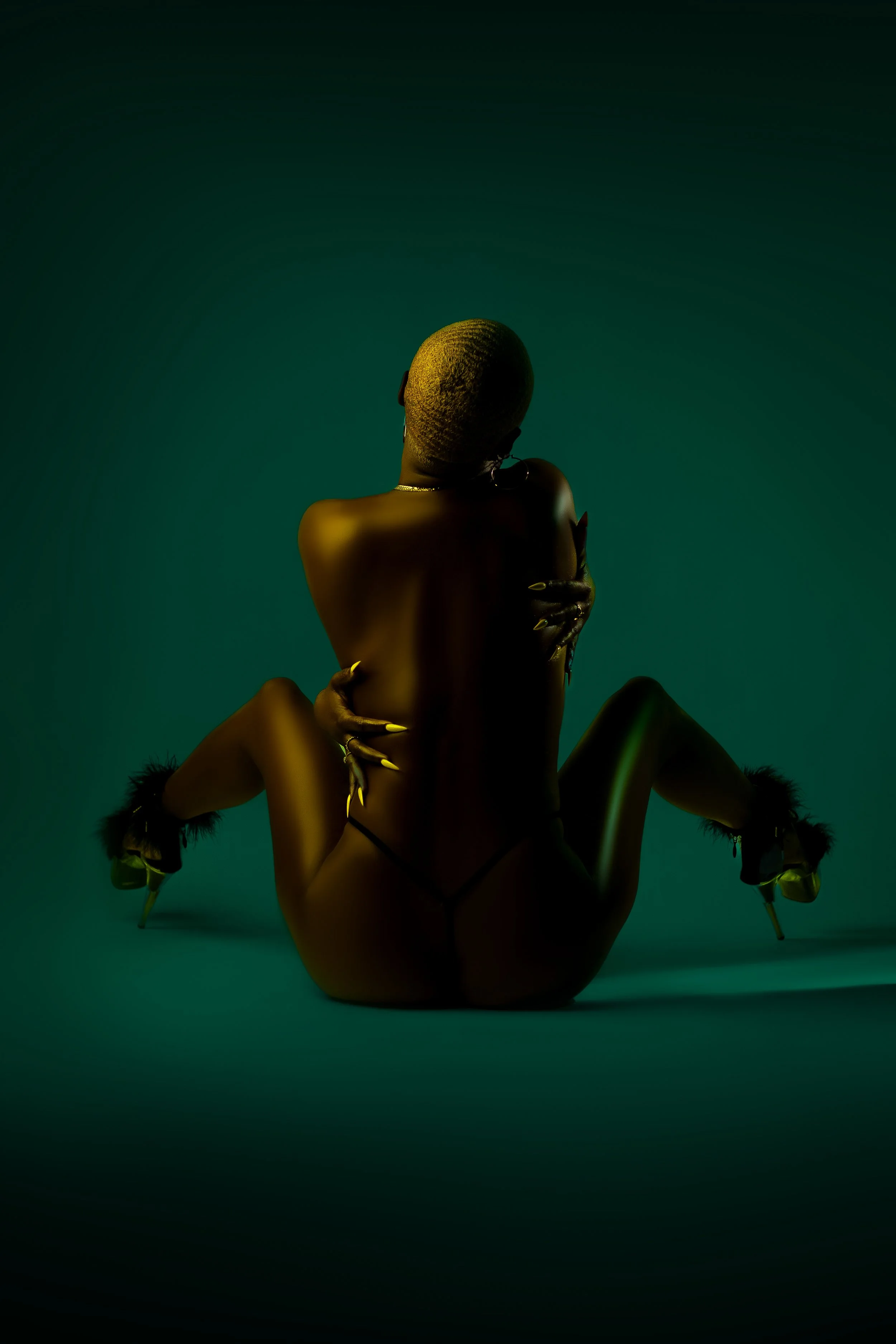 The image shows a nude person with short hair sitting on the ground, facing away from the camera. The person's arms are crossed over their chest, and their head is slightly bowed. The background is a gradient of dark green to black, with soft lighting highlighting the person's back and shoulders.