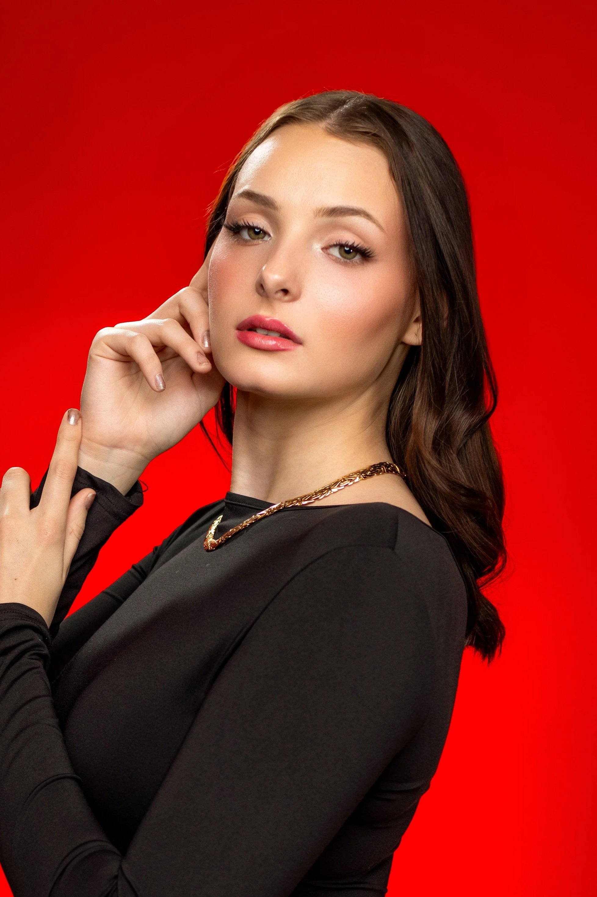 A young woman with long brown hair and fair skin poses against a red background. She wears a black long-sleeve top, gold jewelry, and has makeup with pink lipstick and eyeliner. She is touching her face with her right hand.