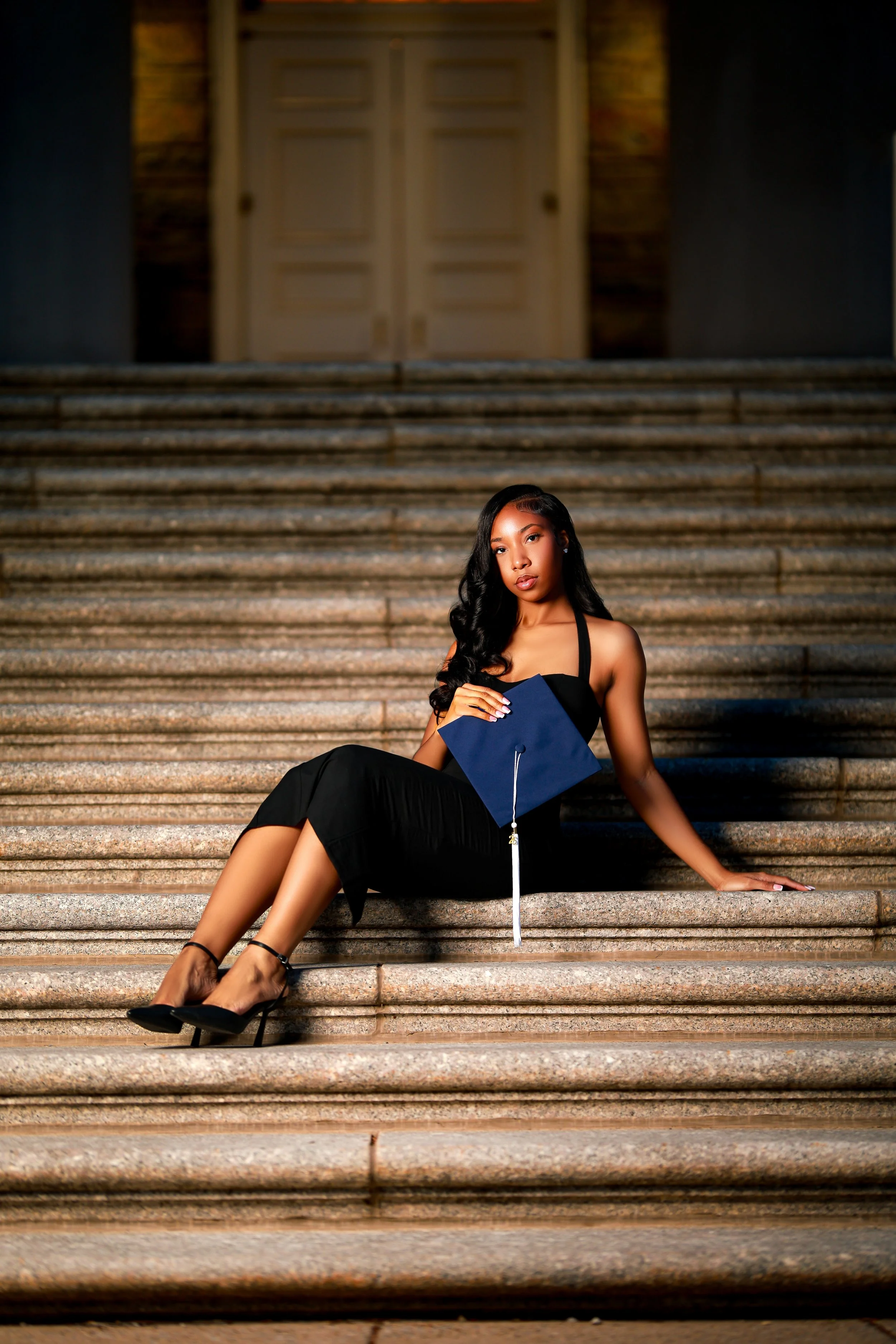 A young woman in a black dress and high heels is sitting on stone steps in front of a large building with double doors. She is holding a blue graduation cap with a white tassel and is looking at the camera.