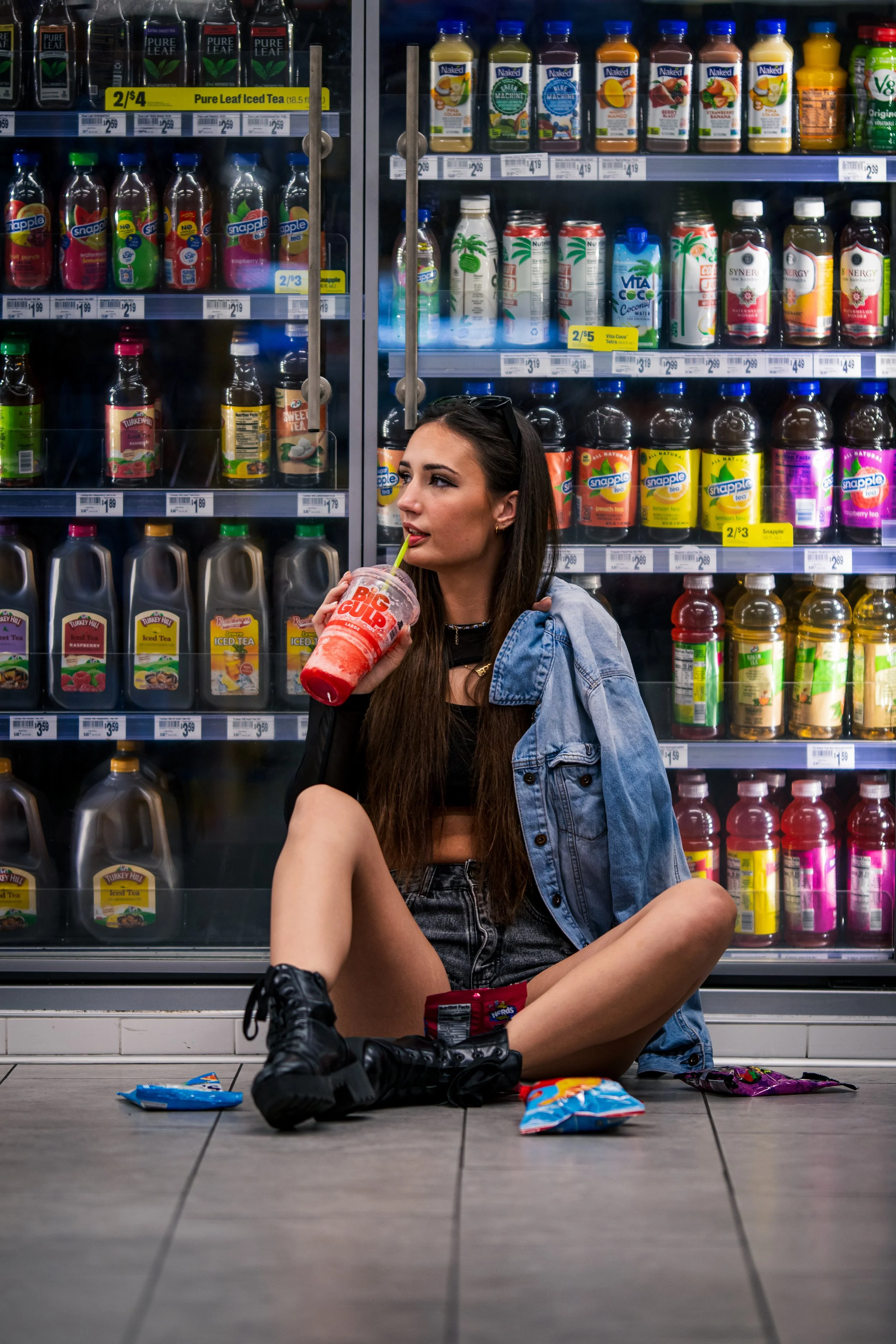 A young woman sitting on the floor of a grocery store aisle, surrounded by snack food and beverages, drinking from a large CrossFit-themed cup with a straw, wearing a black crop top, denim jacket, black combat boots, and black shorts.