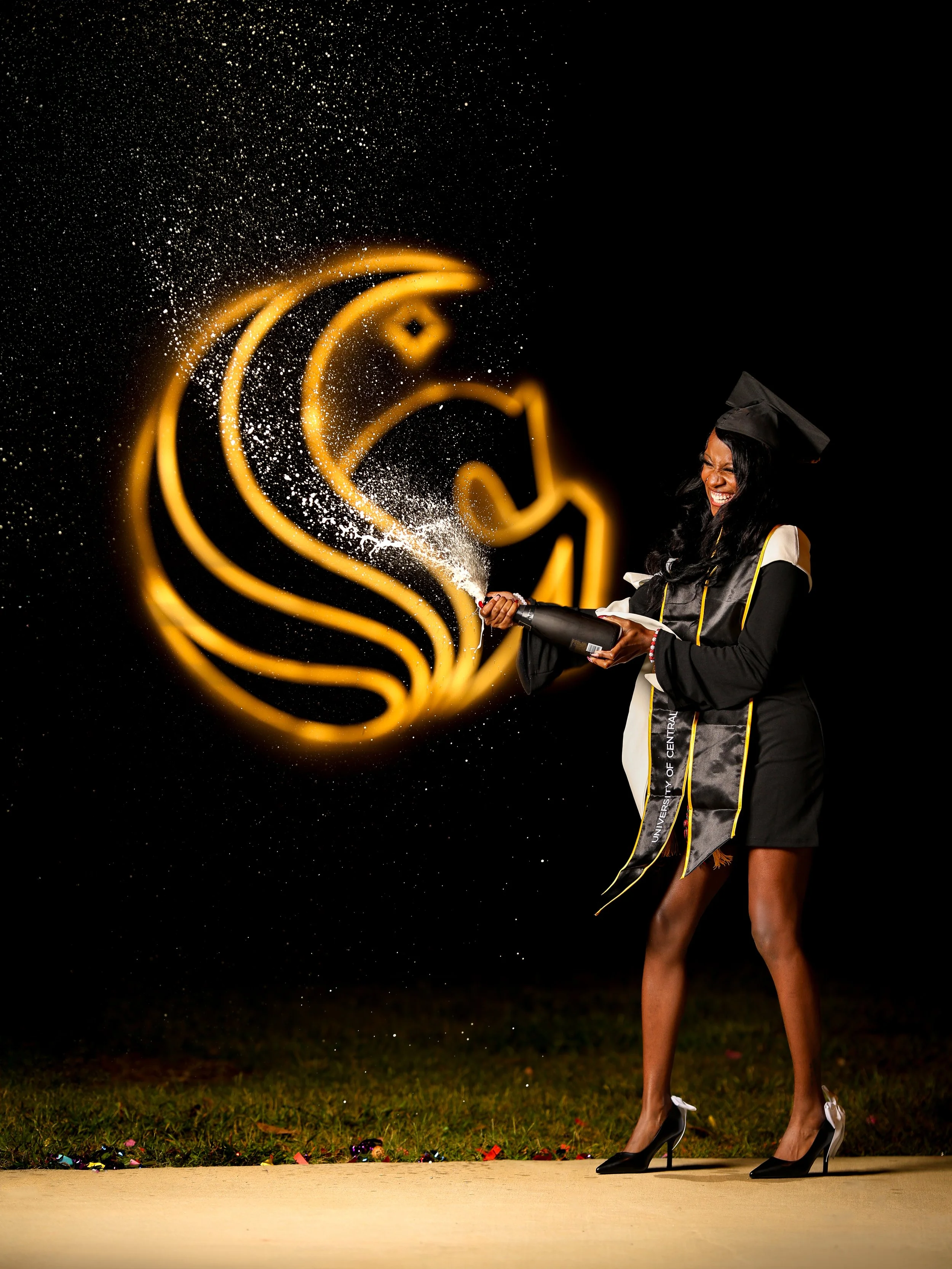 A woman in a graduation cap and gown is standing on a grassy area holding a celebratory confetti popper, which is releasing golden sparks. The background is dark with a glowing, abstract golden symbol or swirl and scattered confetti on the ground.