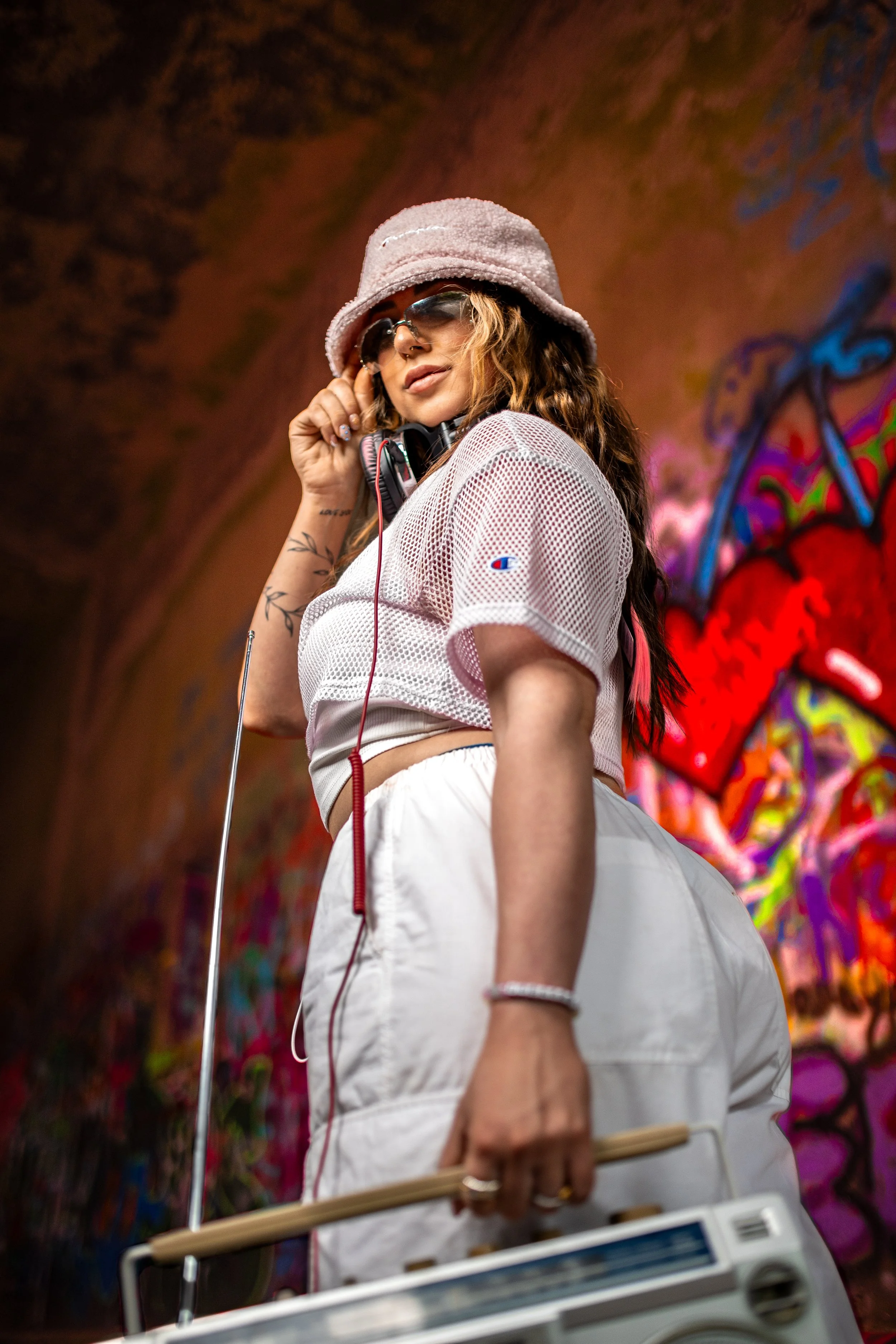 A woman with wavy hair wearing sunglasses and a lavender bucket hat, dressed in a white mesh top and white shorts, holds a water paddle while standing in front of graffiti art in a tunnel.