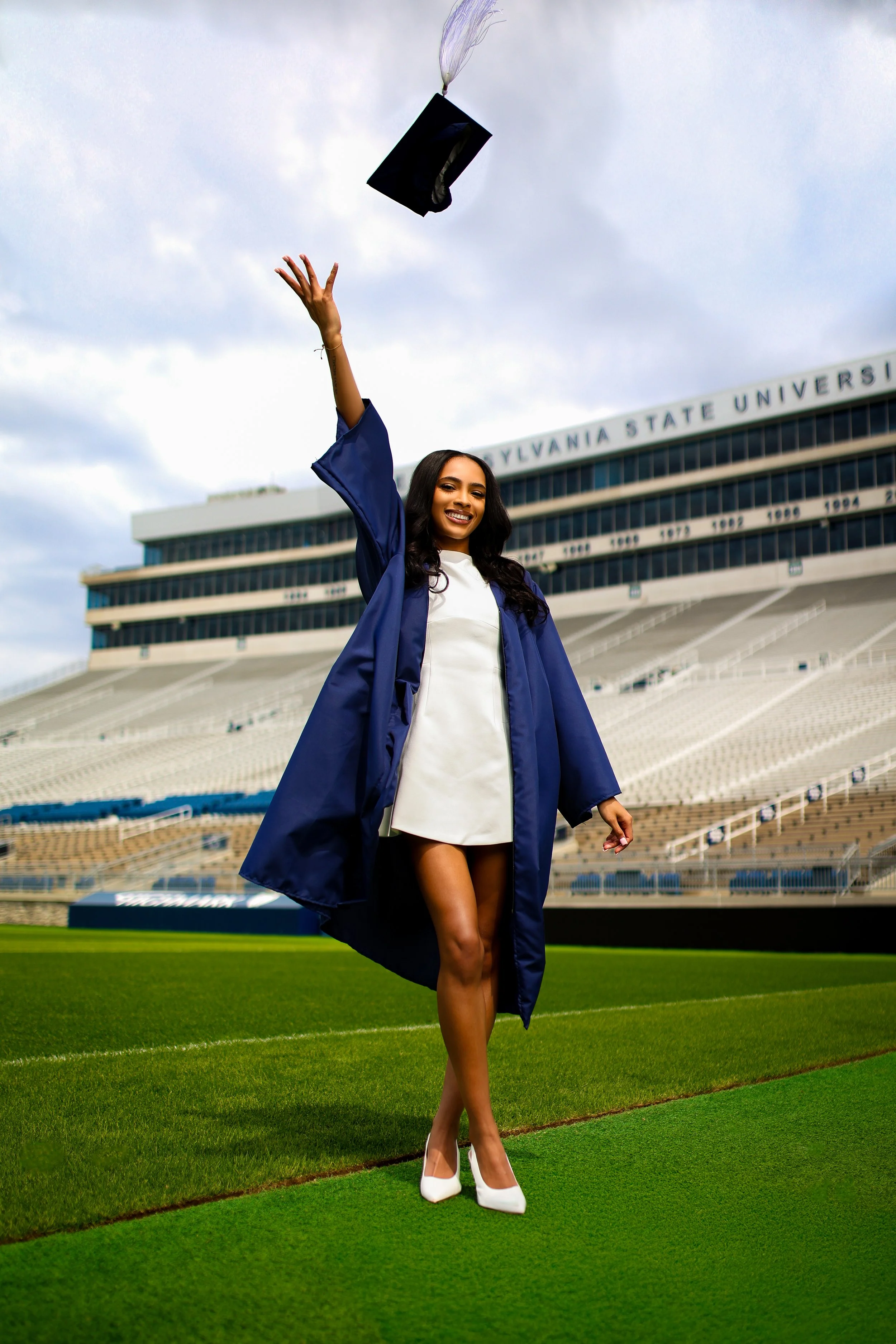 A smiling woman in a graduation gown and cap throwing her cap in the air on a football field with stadium seating and a sign in the background reading "Pennsylvania State University."