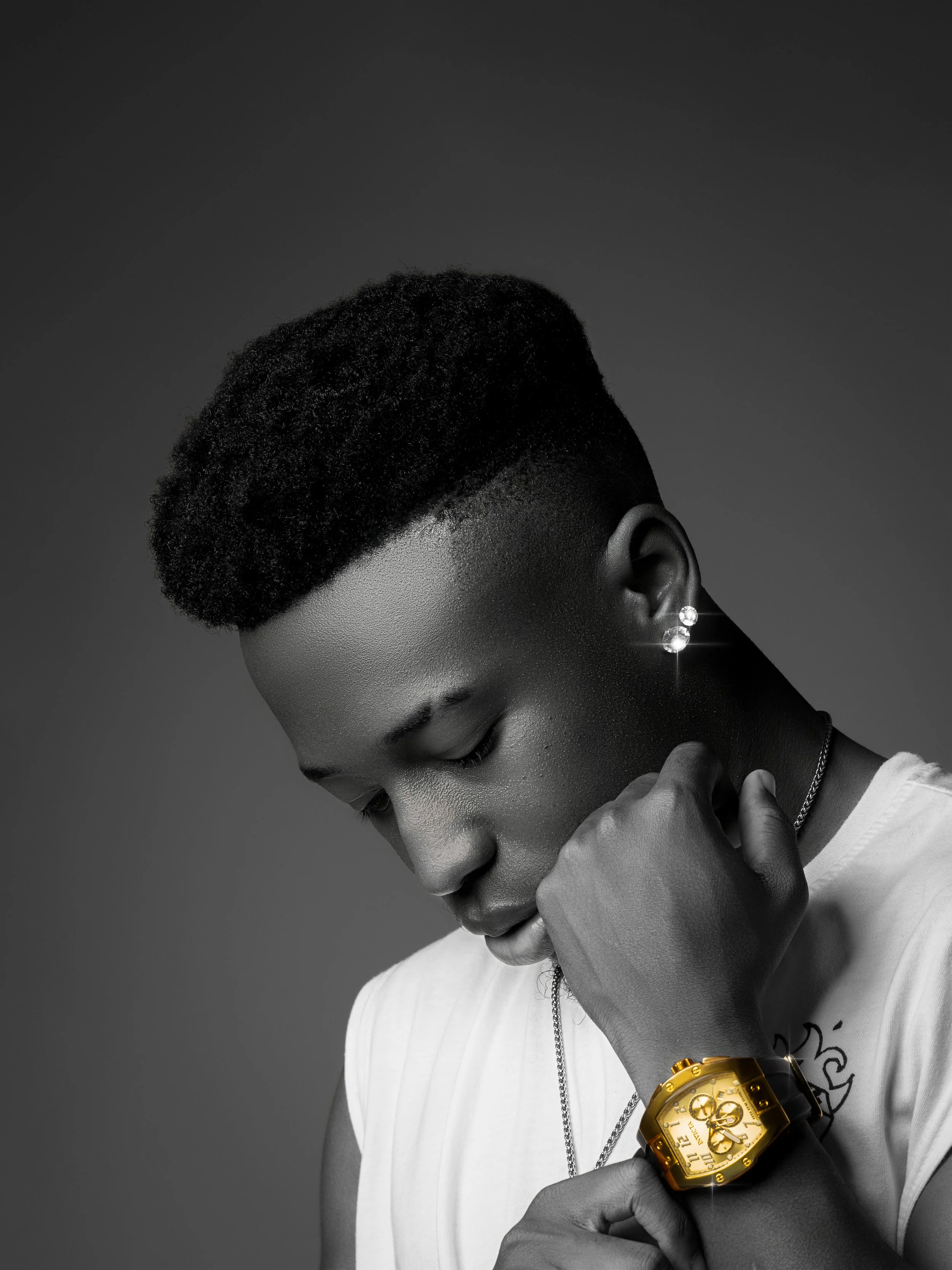 A young woman with a short, textured hairstyle looking down, wearing large earrings, a sleeveless top, and a gold wristwatch with a large face, against a gray background.