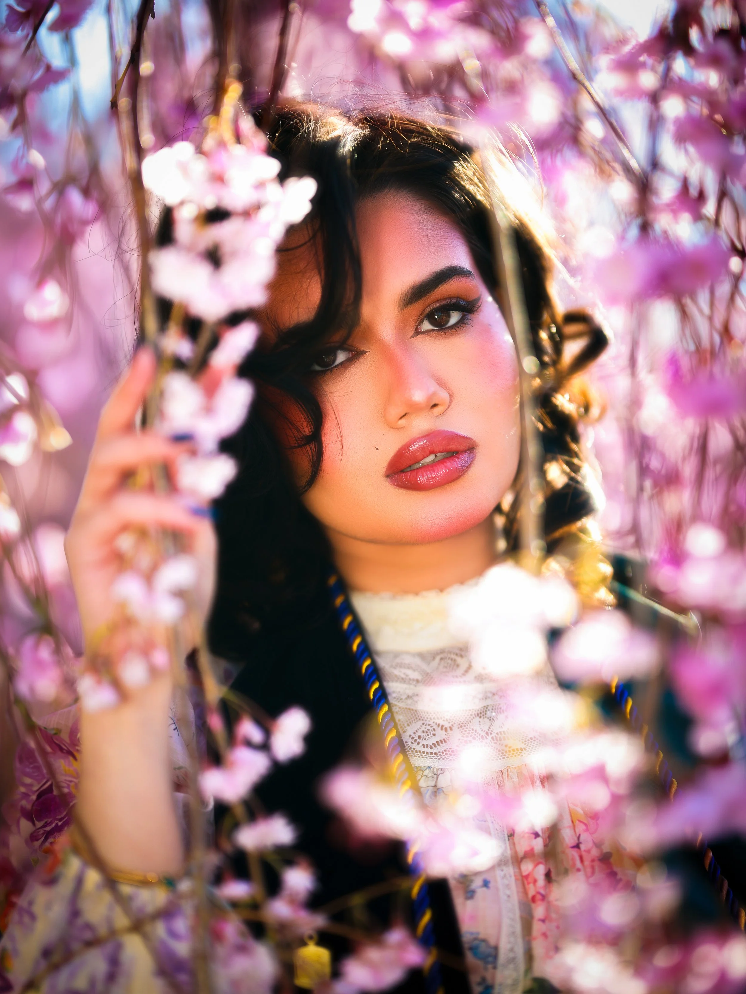 A woman with dark hair and makeup, surrounded by pink cherry blossoms, partially obscuring her face.