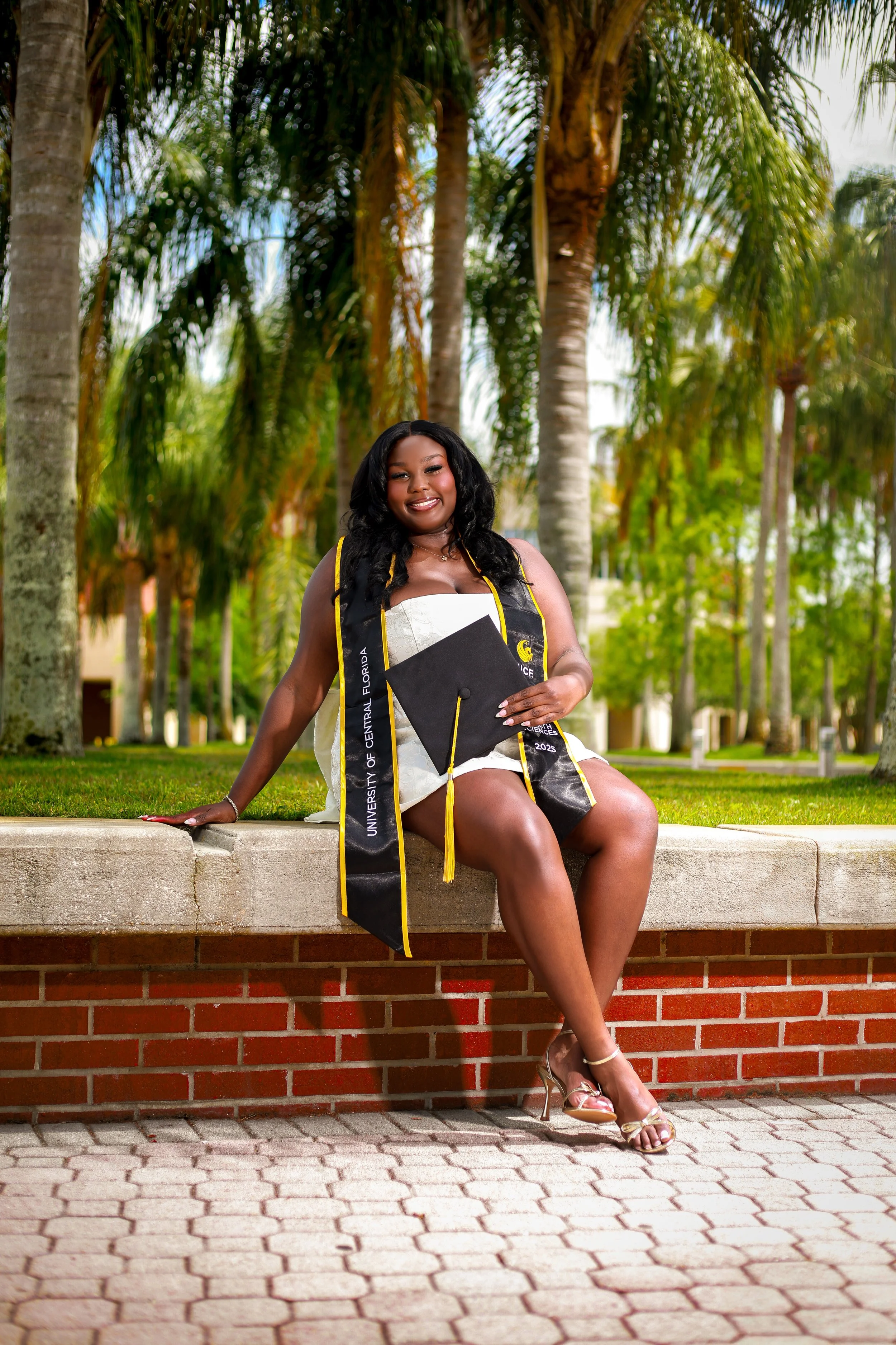 A young woman in a graduation cap and gown sitting outdoors on a stone platform, holding her diploma, with tall palm trees and greenery in the background.