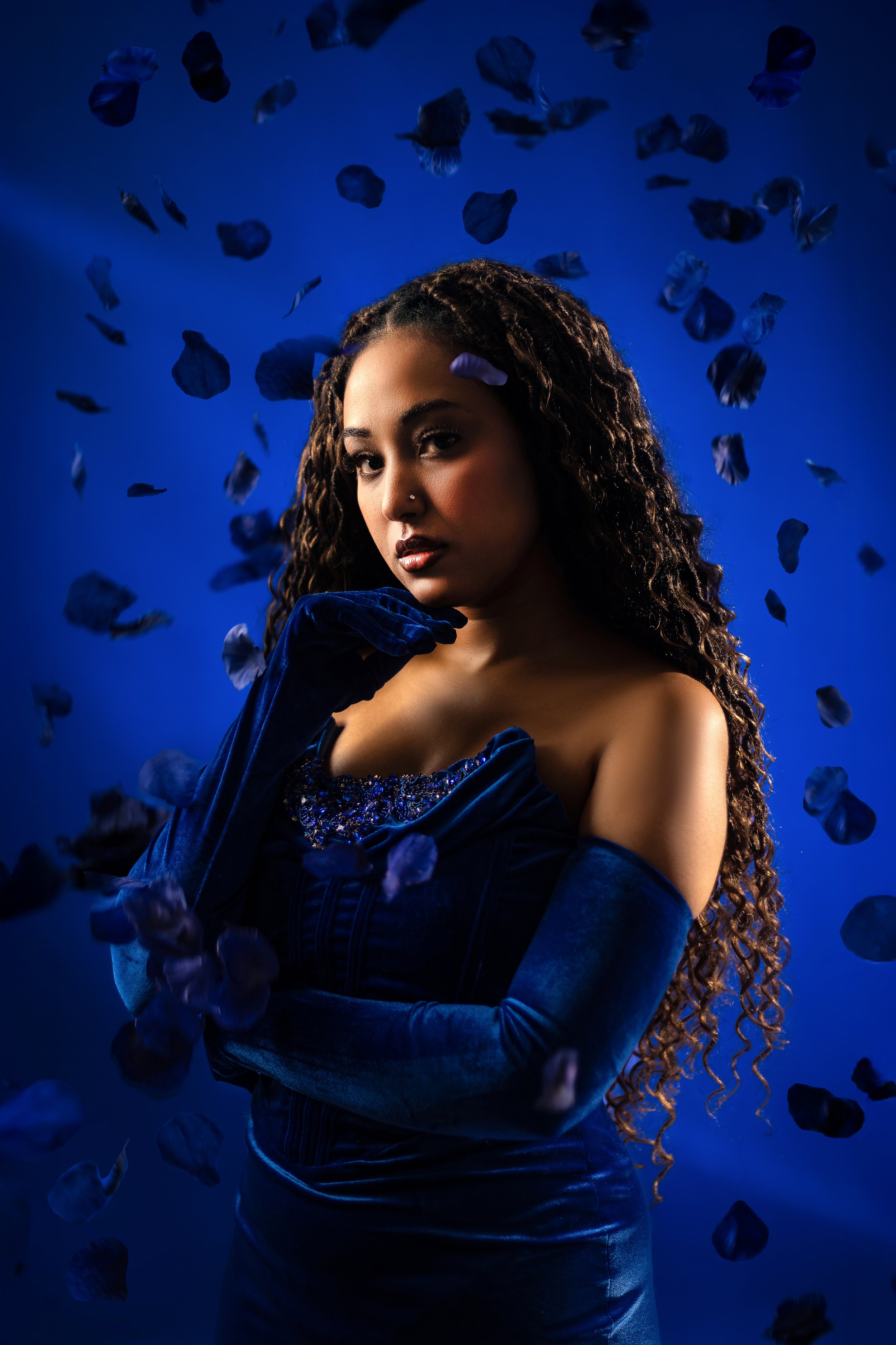 A woman with long curly hair posing against a dark blue background with floating petals, dressed in a dark blue velvet dress and matching gloves.