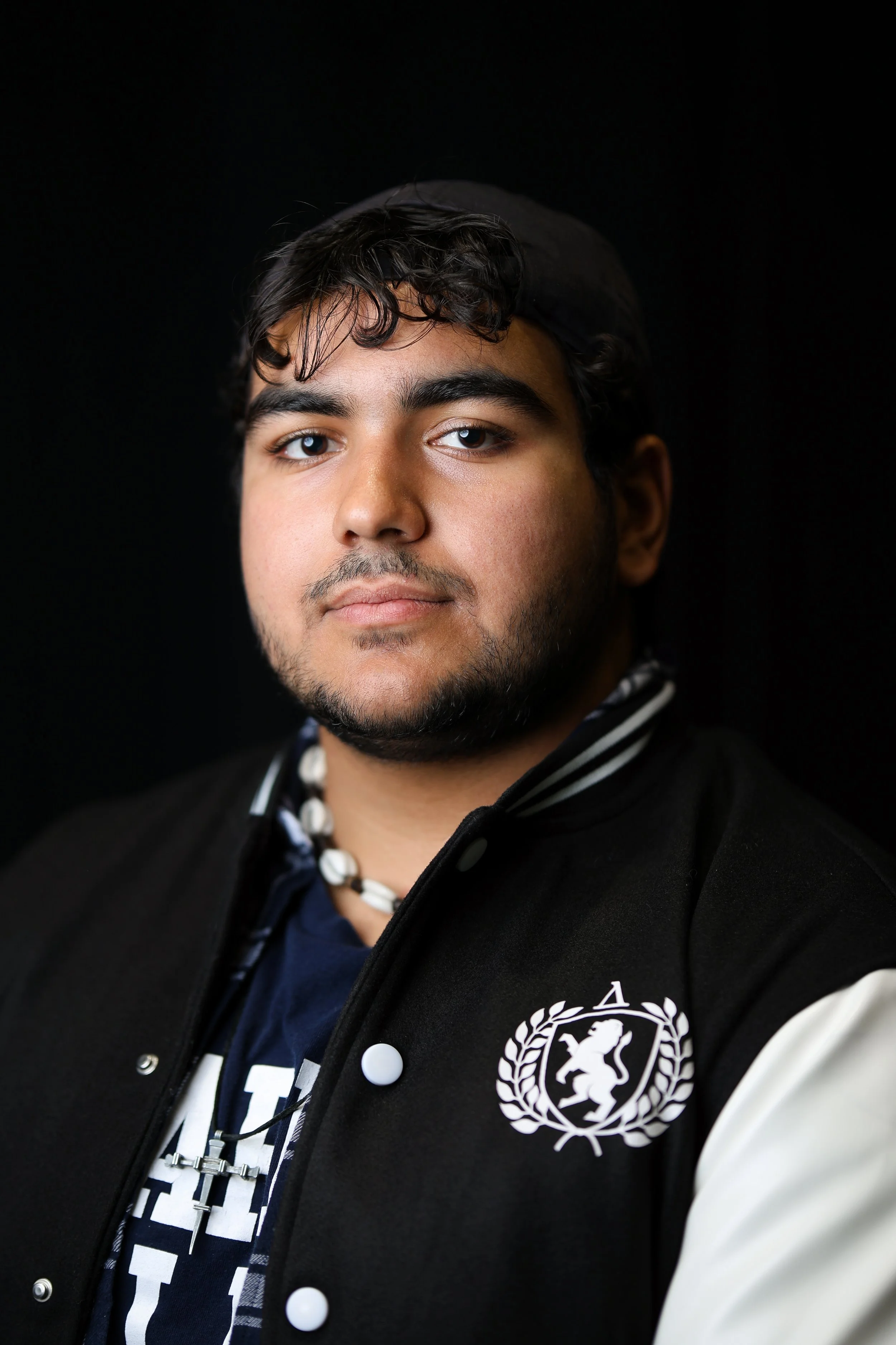 Close-up of a young man with dark, curly hair, beard, and mustache, wearing a black baseball cap backwards, a black and white jacket with a logo featuring a lion, and a black and white striped shirt, against a plain black background.
