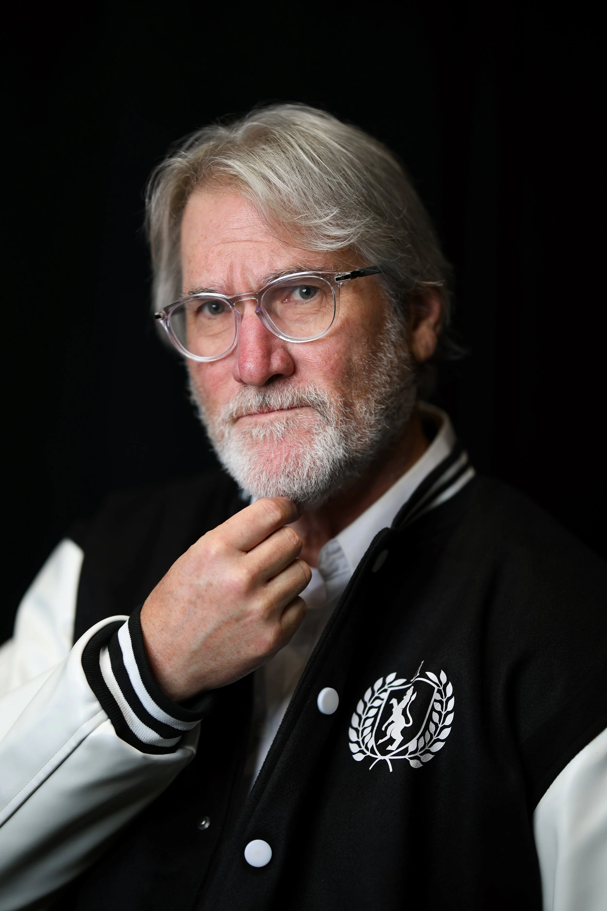 A man with gray hair, glasses, and a beard wears a black and white jacket with a UN logo, with a dark background.