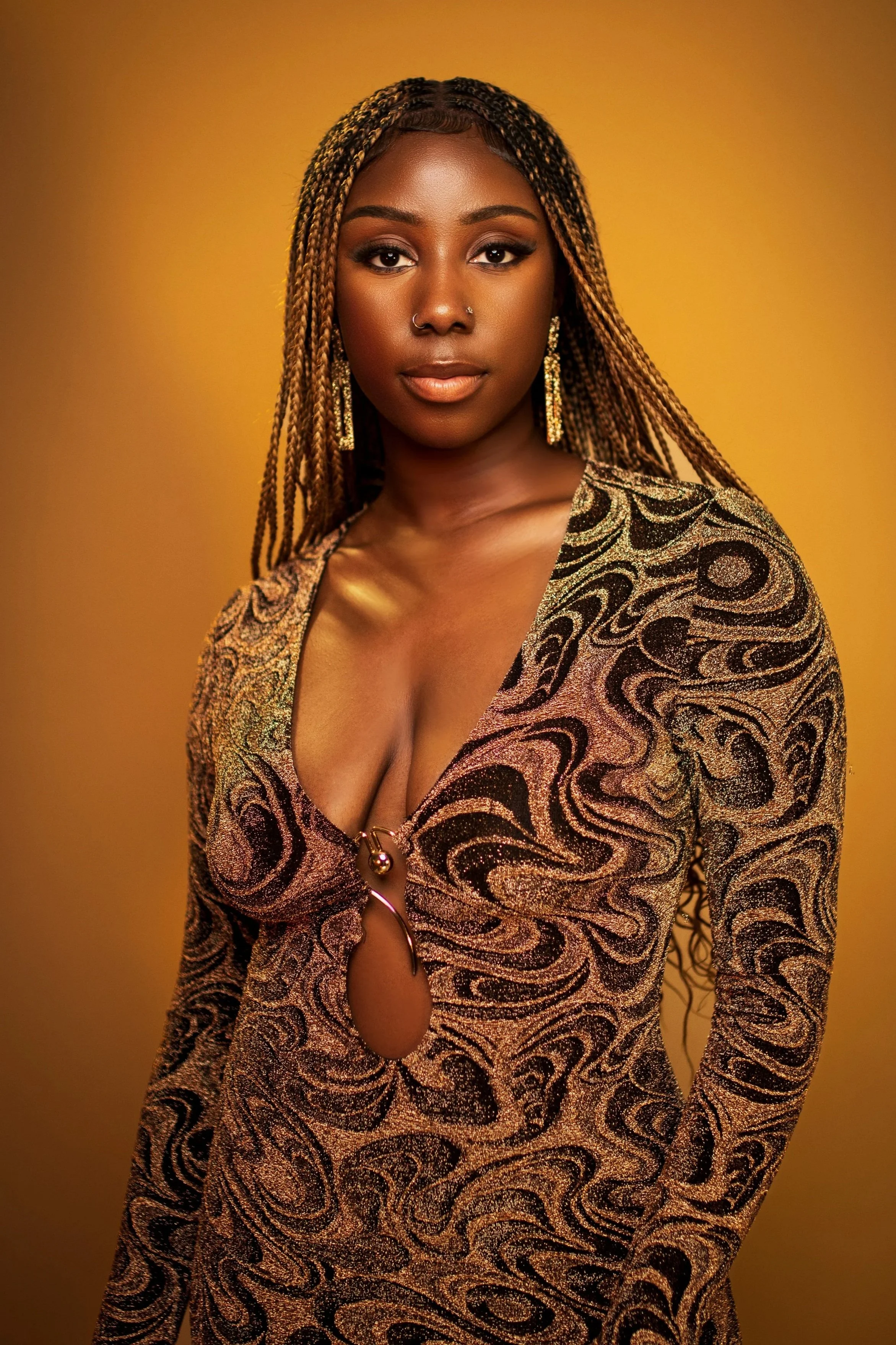 A woman with dark skin, long braided hair, and gold earrings, wearing a gold and black patterned dress with a revealing neckline, against a yellow background.
