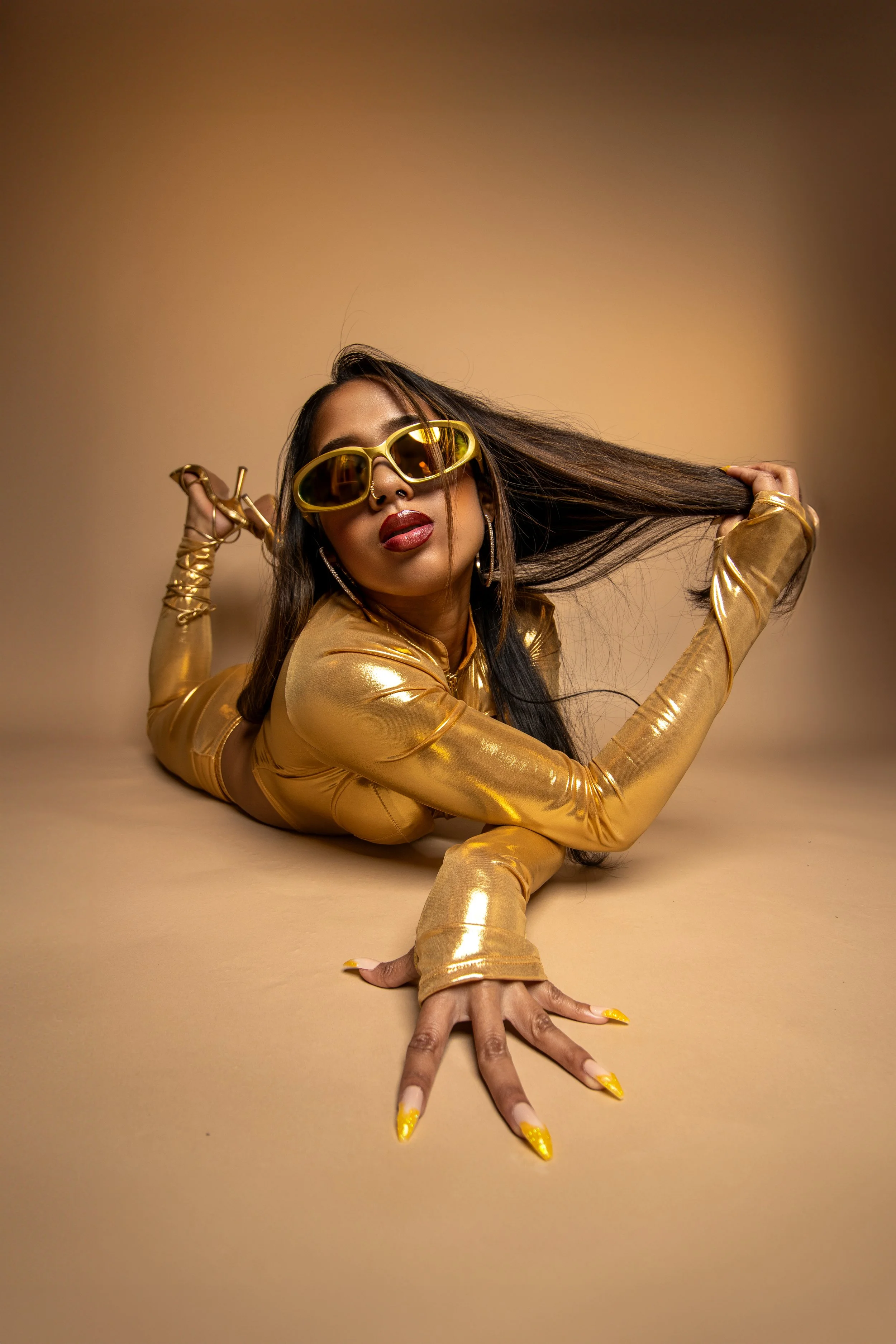 A woman lying on her stomach on a beige surface, wearing a shiny gold outfit and large yellow sunglasses, holding her long dark hair with one hand and stretching her hand forward with the other. She has red lipstick, hoop earrings, and yellow nail polish.