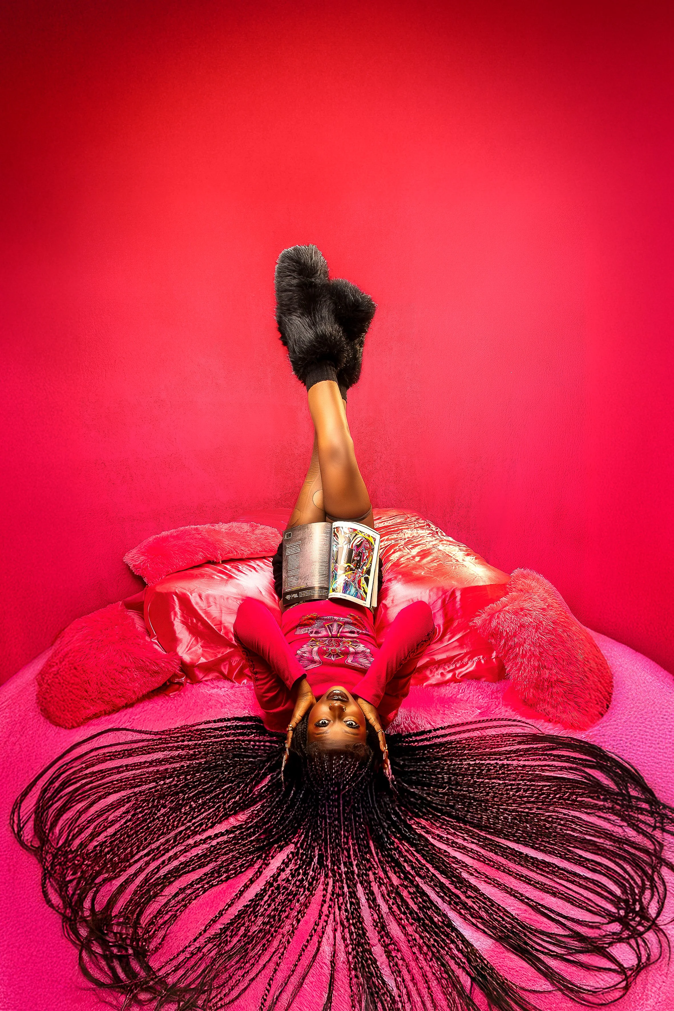 A woman lying on a round pink bed with black hair styled in long braids, against a bright pink wall, wearing a red outfit, holding an open magazine, with a black fuzzy sock on her raised foot.