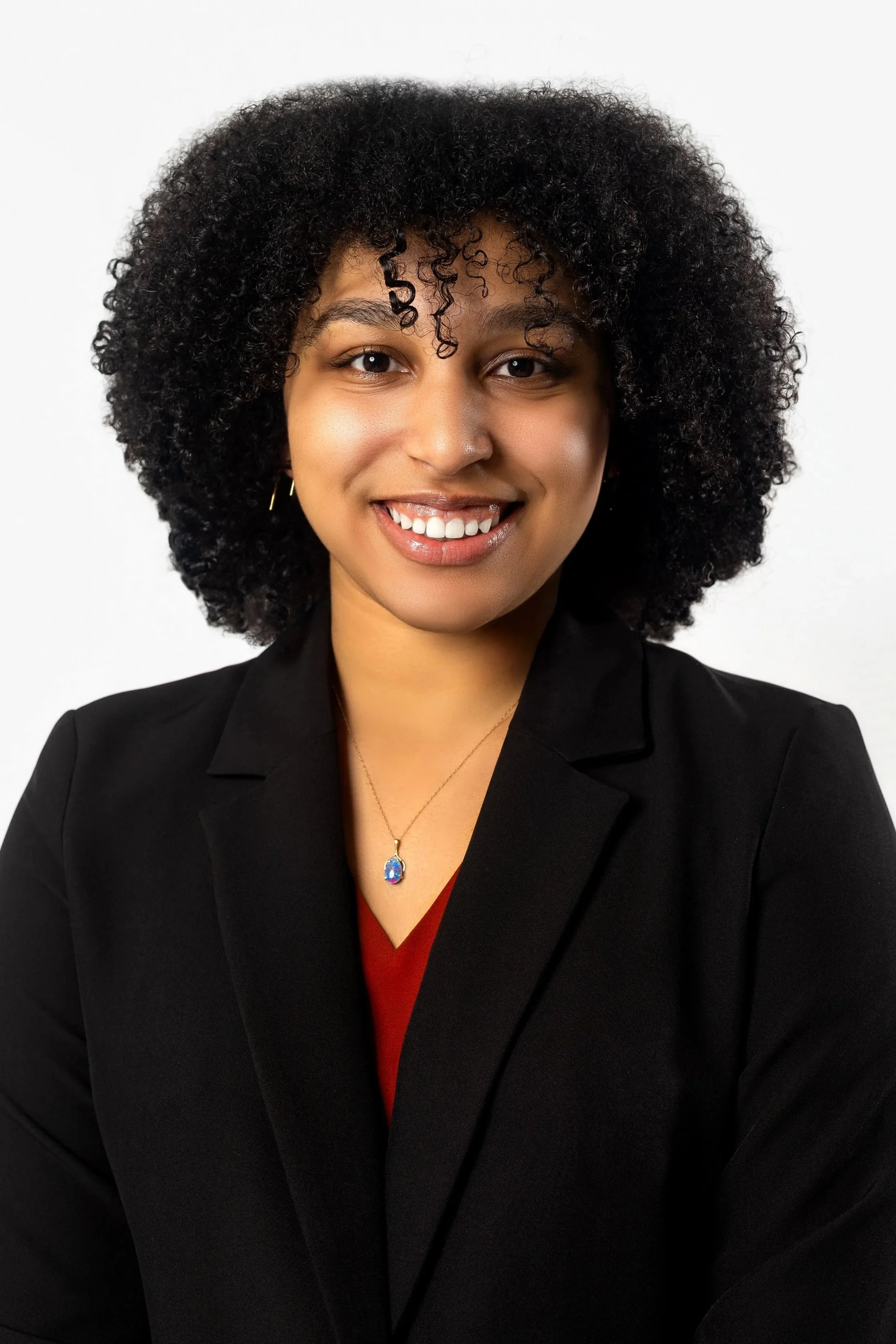 A professional woman with curly black hair, wearing a black blazer, red top, and a necklace with a blue pendant, smiling against a white background.