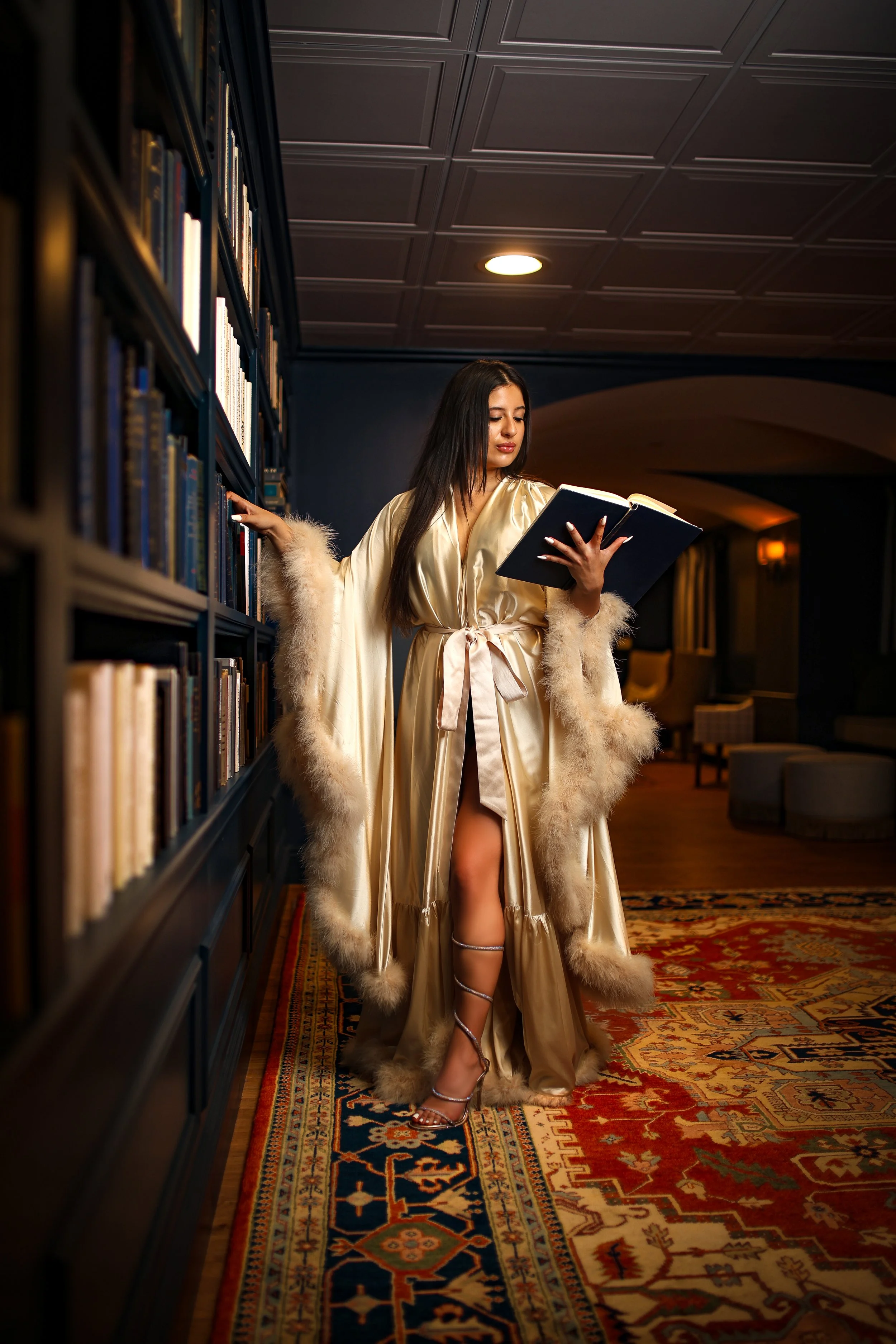 A woman in a luxurious golden gown with fur trim stands in a library aisle, reading a book.