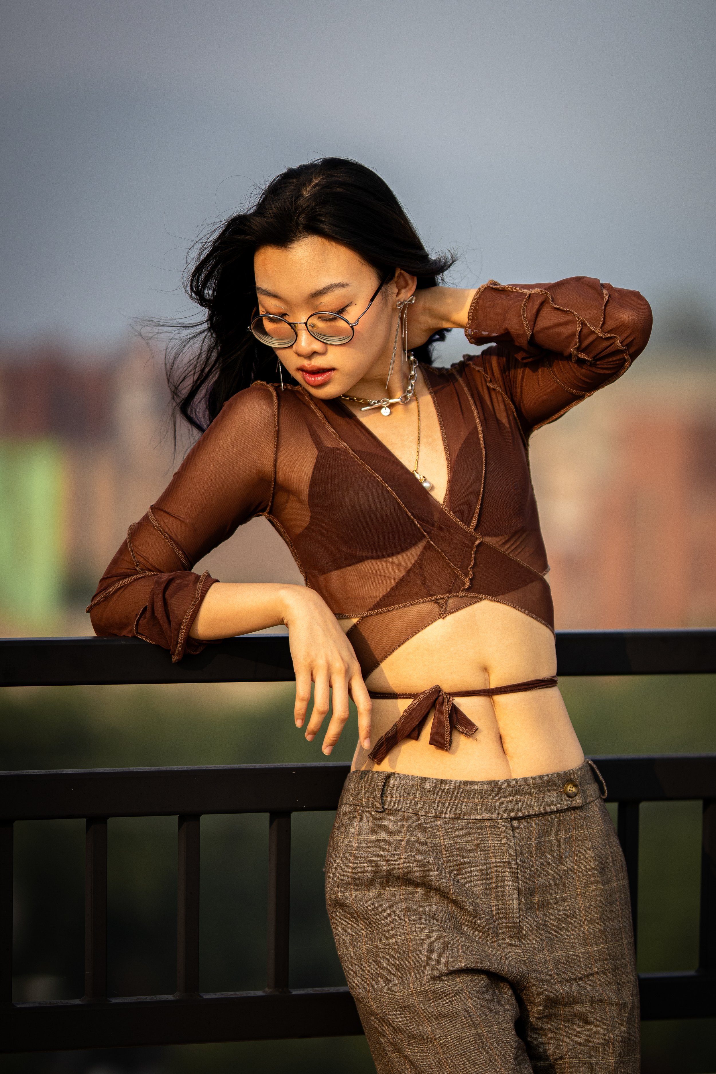 A woman with dark hair, glasses, and layered jewelry leaning on a black railing, wearing a sheer brown top and plaid high-waisted pants, outdoors with a blurred cityscape background.