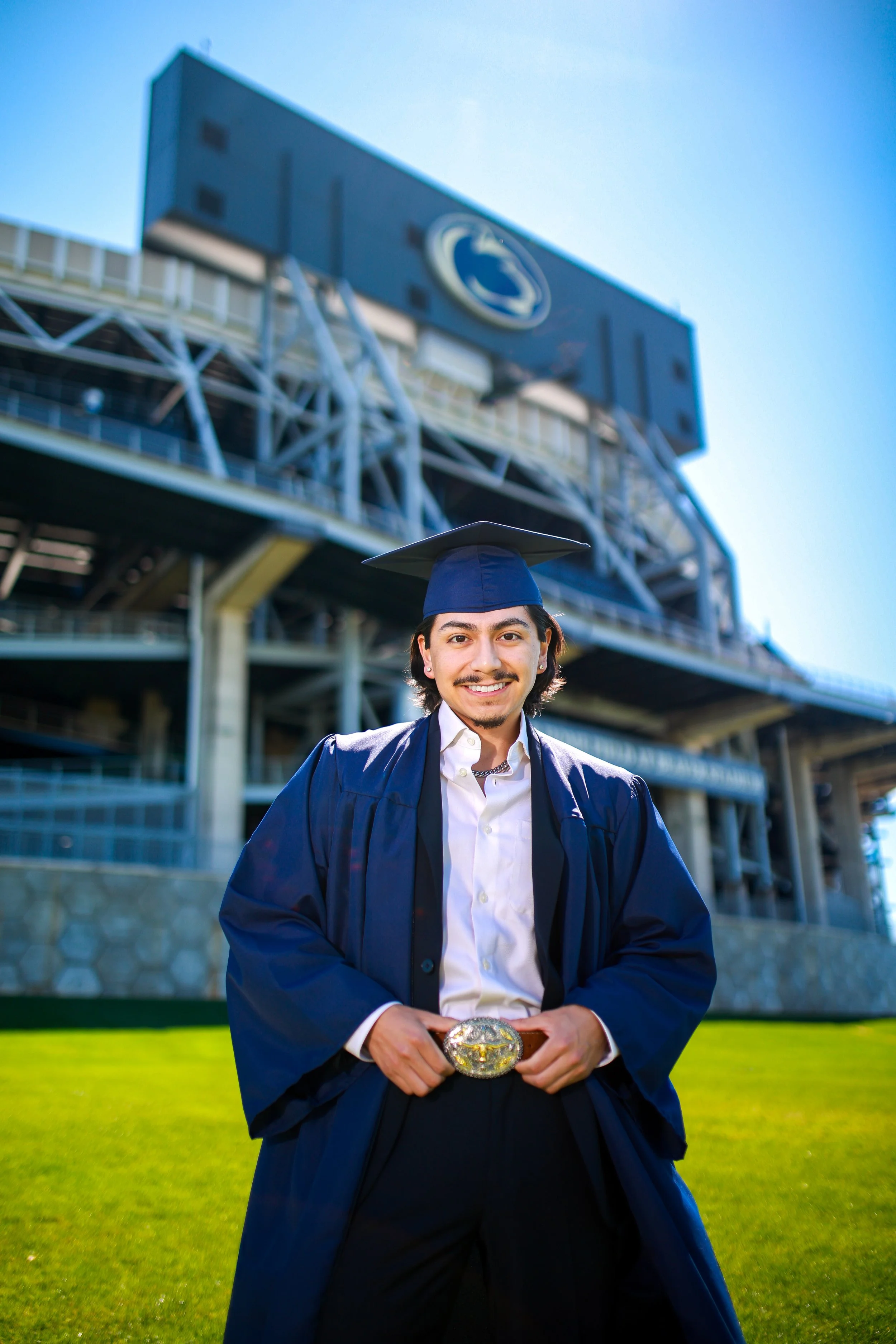 A young man wearing a graduation cap and gown stands outdoors in front of a building with a Penn State logo, holding a rolled diploma with a large ornate belt buckle, smiling at the camera.